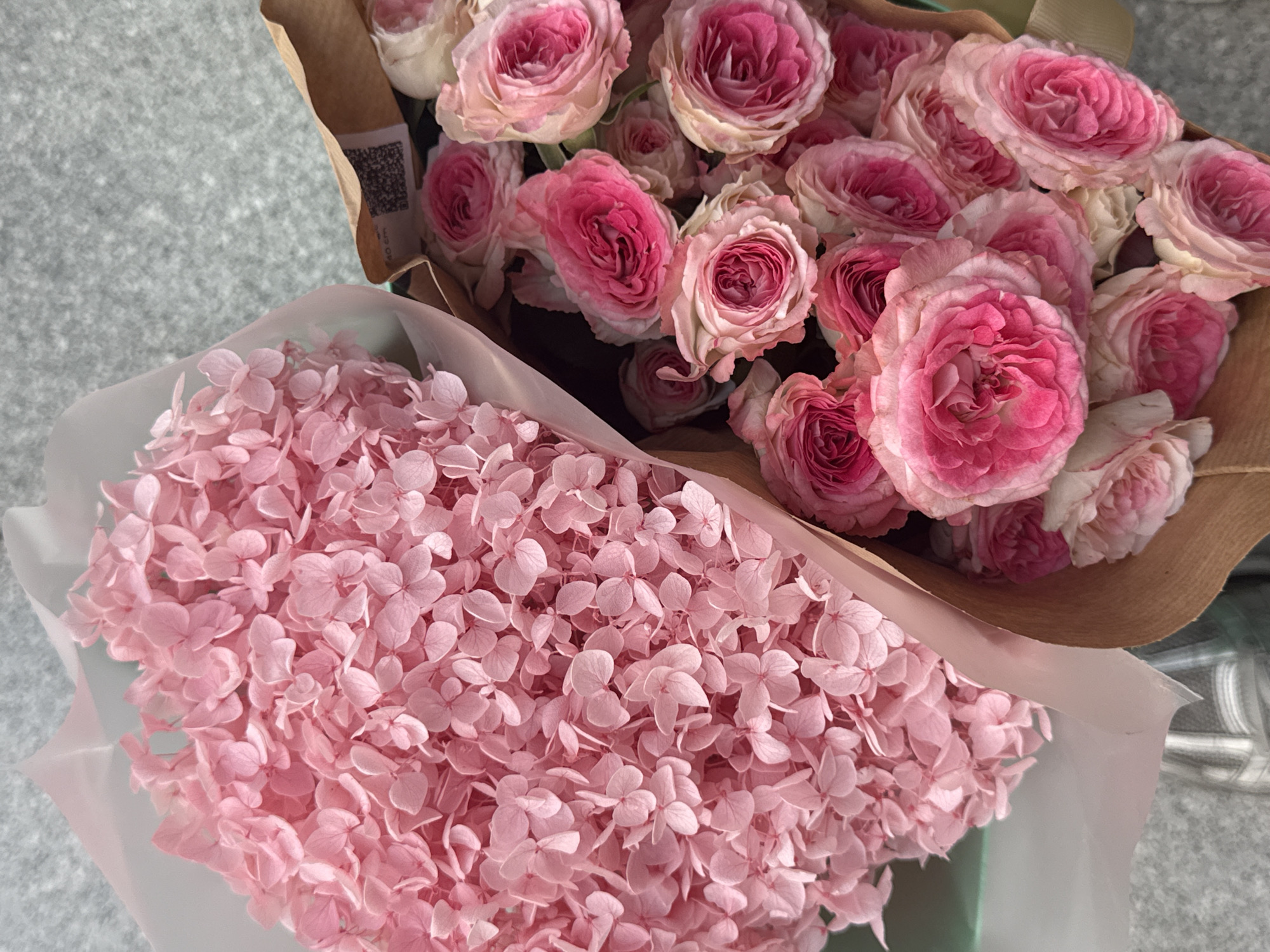 The image depicts two bouquets of flowers, one containing pink roses and the other containing pink hydrangea flowers, placed on a grey surface. The roses are bunched together and wrapped in brown paper, with a QR code visible on the paper. The hydrangea bouquet is wrapped in light pink translucent paper. The location is not discernible from the image and there is no indication that it is in Dubai, United Arab Emirates. No people are visible.