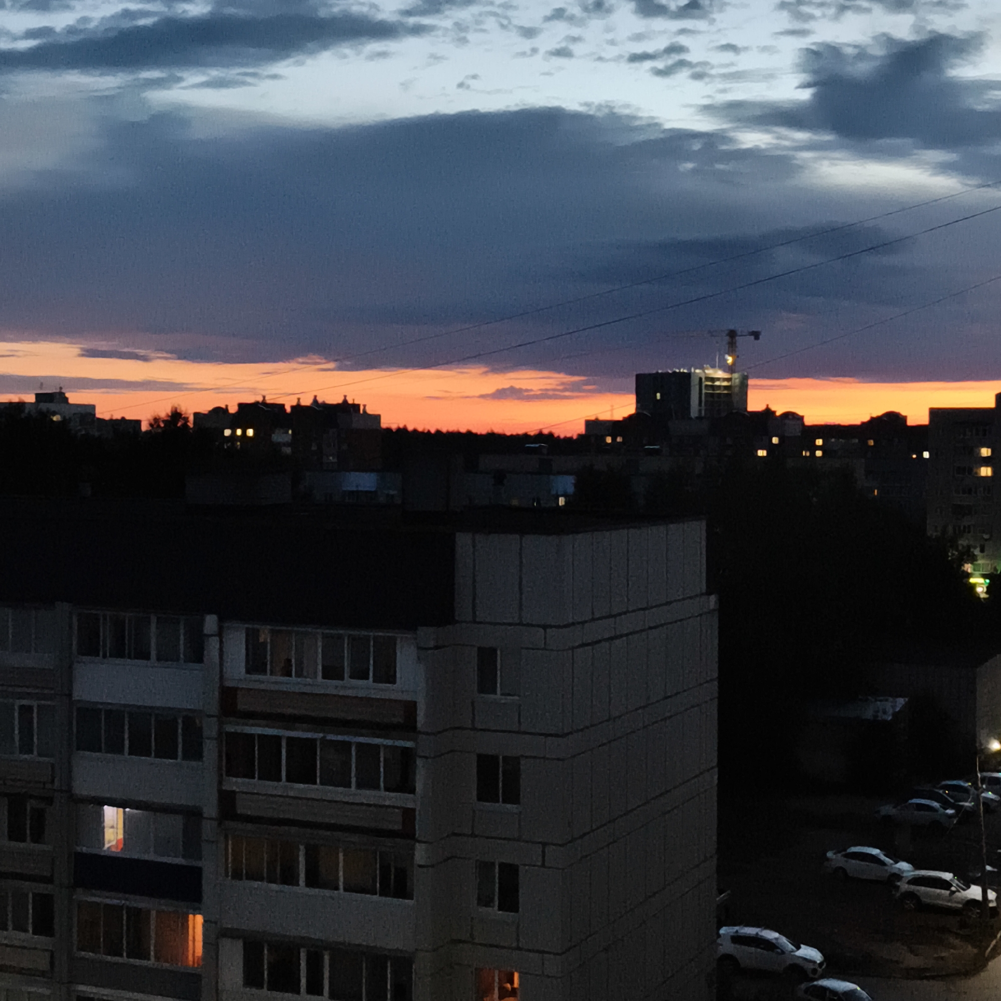 The image depicts a twilight scene in the city of Chaikovsky, Russia. In the foreground, the side of a multi-story residential building is visible, featuring numerous windows, some of which are illuminated with warm, orange light. The building's facade is constructed of concrete panels. Beyond this building, the cityscape unfolds with other structures, including apartment buildings and what appears to be a construction site with a crane. The sky above is a gradient of deep blue and orange, indicating the setting or rising sun, with scattered clouds. Power lines are also visible stretched across the sky. In the lower right portion of the image, a dark asphalt area shows several parked cars, predominantly white, suggesting a residential parking lot. The overall atmosphere is one of evening transitioning into night in a Russian urban environment.