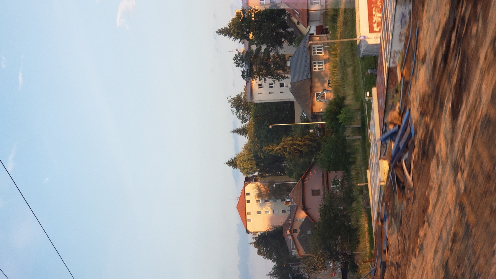 The image shows a suburban area in Poprad, Slovakia, bathed in the warm light of the setting sun. In the background, several multi-story buildings and houses are visible, nestled amongst lush green trees. One prominent building, painted in a light yellow hue with a red roof, stands out. In the foreground, a construction site is evident, with piles of lumber, pipes, and building materials scattered across the dusty ground. A few individuals can be seen near the construction area, suggesting ongoing work. The sky is clear with a few wispy clouds, indicating fair weather. The overall scene depicts a blend of residential life and ongoing development in a European town.