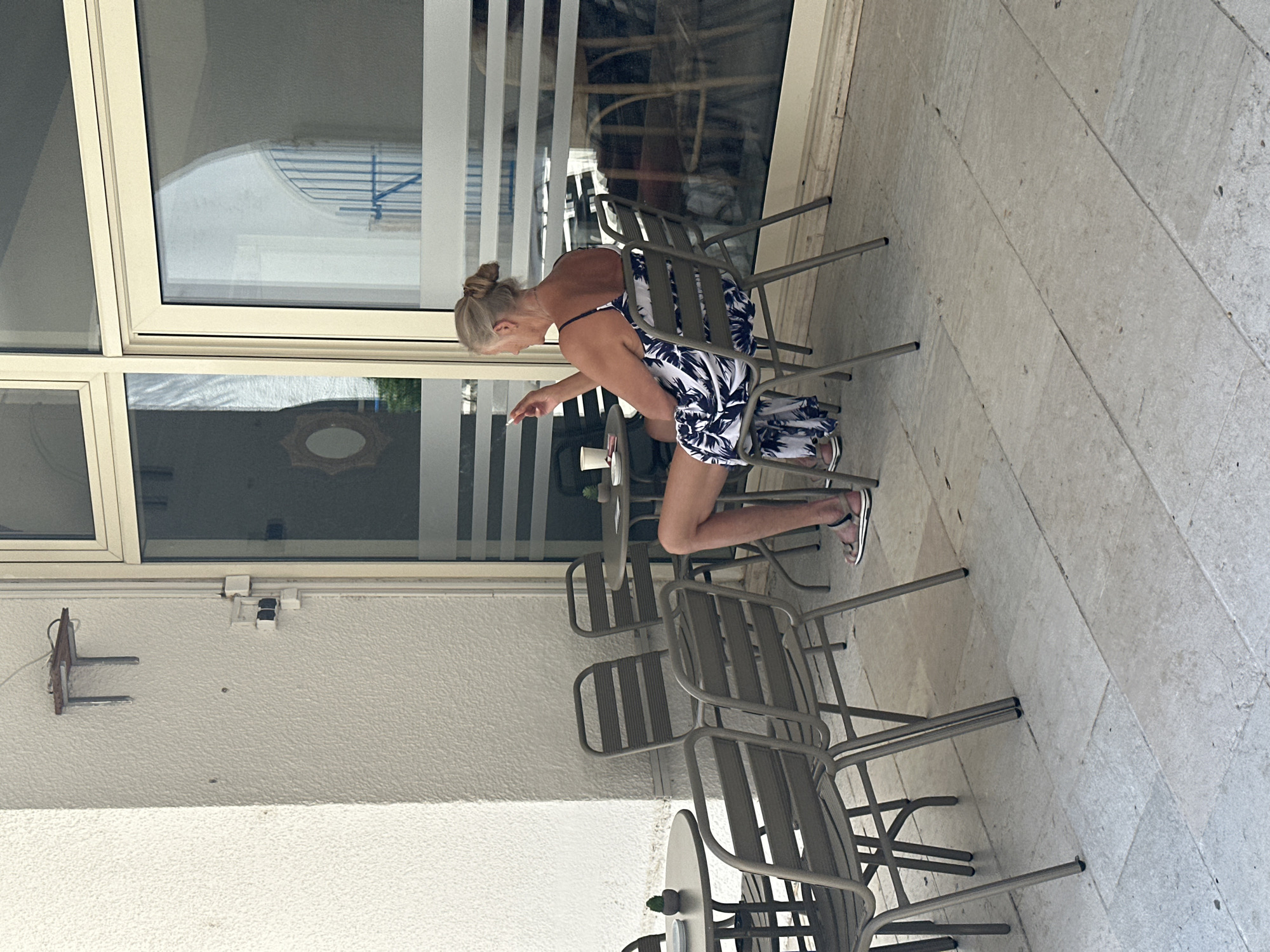A woman with blonde hair pulled up in a bun sits outdoors at a cafe table in Budva, Montenegro. She is wearing a sleeveless blue and white floral dress and white sandals. She is holding a lit cigarette in her right hand and appears to be smoking. The woman is seated in a metal chair with horizontal slats. In front of her, on a round metal table, is a white cup and saucer. The cafe has a paved stone floor and is adjacent to a building with white textured walls and large glass windows with horizontal blinds. Several other empty metal chairs and tables are visible in the foreground and background. The reflection in the windows shows an arched entryway and scaffolding.