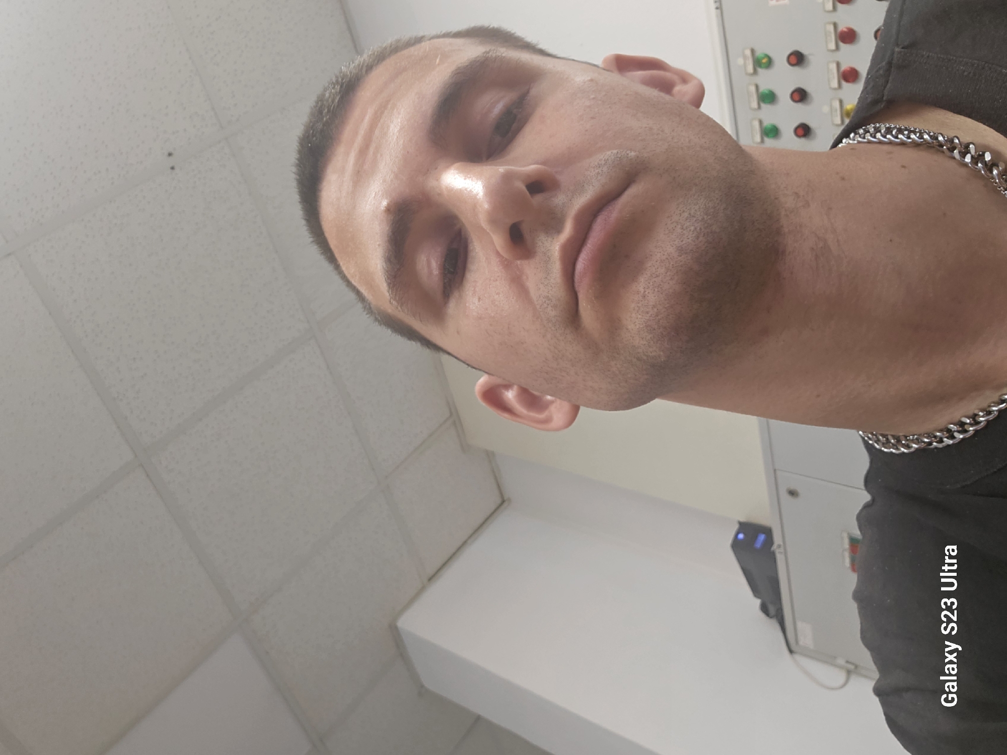 The image is a selfie of a man taken indoors. He has short brown hair and is looking down at the camera with a neutral expression. He is wearing a black t-shirt and a silver chain necklace. In the background, there is a ceiling with acoustic tiles, white walls, and some electrical equipment. The text "Galaxy S23 Ultra" is overlaid in the lower left corner, indicating the device used to take the photo. There is no visible indication of the specific location within the city of Haskovo, Bulgaria.