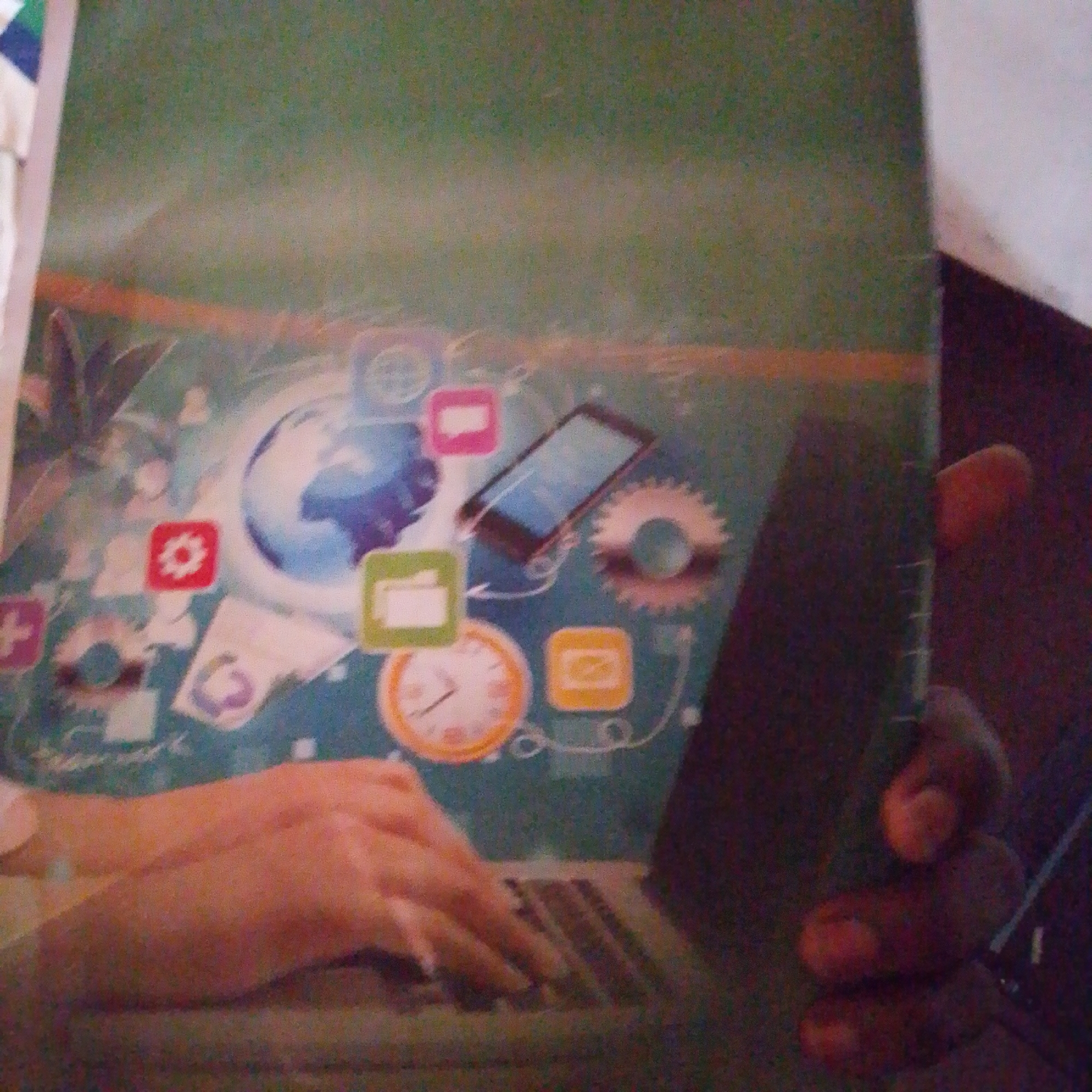 The image is a close-up, angled shot of what appears to be the cover of a book or magazine, featuring a digital-themed illustration. The illustration depicts a globe surrounded by various app icons, a smartphone, and gears, suggesting themes of technology and connectivity. In the foreground, a person's hands are visible, with one hand resting on a laptop keyboard, implying engagement with technology. The lighting is dim, and the image quality is grainy, making specific details difficult to discern. There is no discernible information within the image that would allow for the identification of its location in Jalingo, Nigeria.