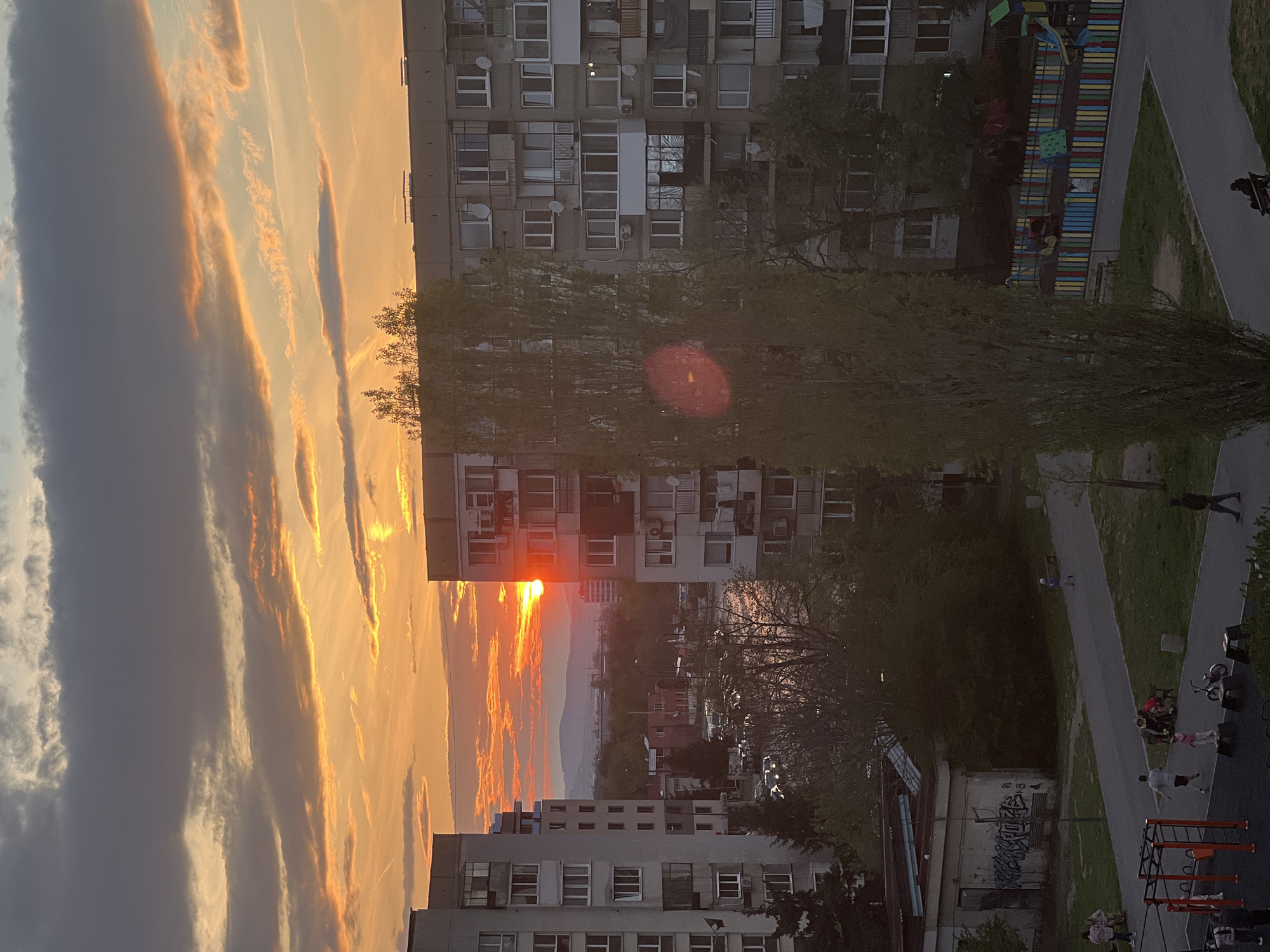The image shows a cityscape at sunset, likely taken from a high vantage point in Sofia, Bulgaria.
**Environment:** A dramatic sunset sky dominates the left side of the image, with streaks of orange and yellow light interspersed with grey and white clouds. The setting sun is visible behind a high-rise building in the middle ground. The buildings appear to be typical residential structures, possibly built during the communist era. A park and playground area is visible to the right of the buildings with people visible there.
**Buildings:** The buildings are typical post-soviet era residential. A lot of windows, several stories and some wear and tear on the exterior.
**People:** Several people can be seen interacting in the playground.
**Objects:** Trees in the park and the playground structure can be seen.