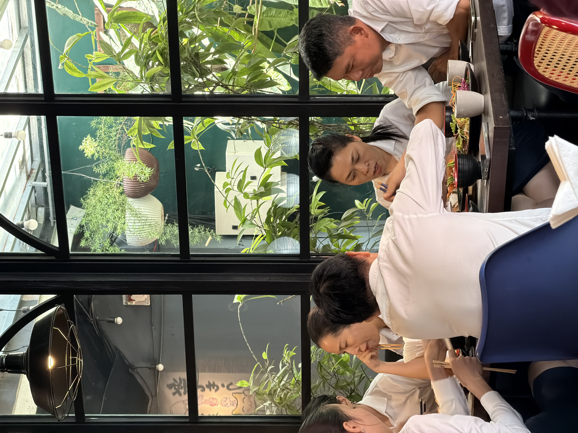 A group of four individuals, two men and two women, are seated at a dark wooden table in a restaurant, engaged in a meal. The scene is viewed from a slightly elevated perspective, looking down at the table and the people. The individuals are dressed in white shirts, suggesting a uniform or a smart casual dress code.
In the foreground, to the left, a woman's arm is visible, holding chopsticks over a plate of food, likely sushi or sashimi, which is partially obscured. Her white shirt sleeve is prominently displayed. To her right, a woman with her hair pulled back is looking down, also holding chopsticks, and appears to be in conversation or discussion with the people around her. Her right hand is resting on her chin.
In the center of the image, a man's back is visible, facing away from the camera, wearing a white shirt. He is seated at the table, and his presence suggests he is part of the group.
To the right of the woman in the center, a man in a white shirt is looking towards the woman, his expression suggesting he is listening or engaged in conversation. His hands are resting on the table.
The table is laden with various dishes, including small bowls of food, plates with what appears to be sushi or rolls, and cups. A smartphone is visible on the table in front of the woman in the center. The background is dominated by large windows with black frames, revealing an indoor garden or courtyard with lush green plants and hanging potted plants. The natural light filtering through the plants creates a serene ambiance. The interior design suggests a modern, possibly Asian-inspired, restaurant. The restaurant appears to be located in Nha Trang Ward, Vietnam, given the context of the prompt.