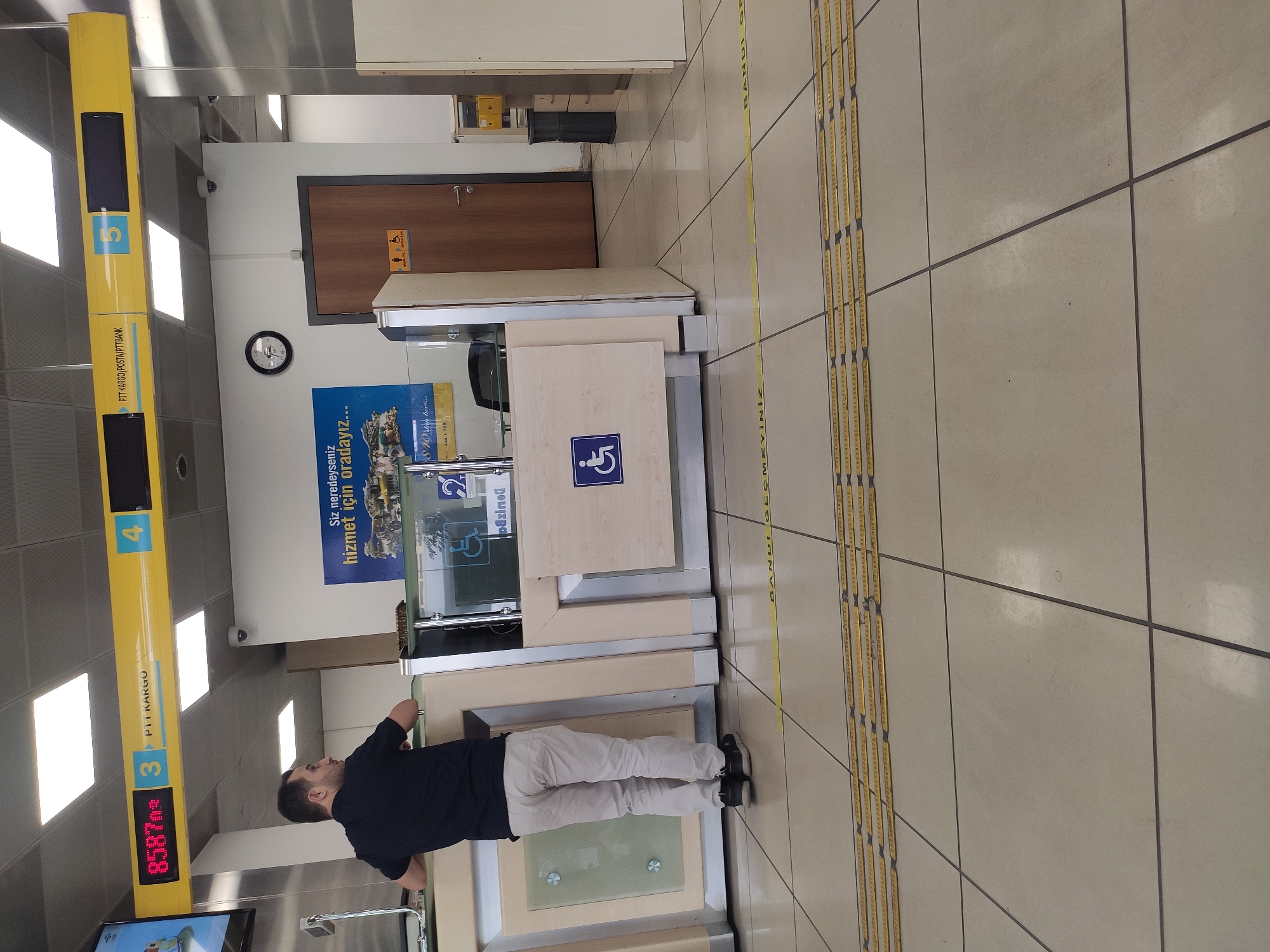 This is a photograph taken indoors, likely in a public service building such as a post office or bank in Ankara, Turkey. The scene is viewed from a slightly elevated angle, looking down and forward. In the foreground, the floor is tiled with large, light-colored square tiles. Yellow tactile warning strips are laid out in parallel rows on the floor, indicating a pedestrian path.
The midground features a counter area with light-colored wood paneling and glass partitions. A man, seen from behind and the side, is standing at the counter, wearing a dark blue t-shirt and light beige trousers. He is looking to his right. To the right of the man, a wooden door is visible, with signs above it indicating a restroom facility for men and women and an accessible restroom. A circular clock with Roman numerals is mounted on the wall above the door. Further to the right, a black trash can and a filing cabinet with yellow drawers are visible.
In the background, a yellow overhead sign displays "PTT KARGO/POSTA/PTTBANK" and numbers "3", "4", and "5" in blue boxes, indicating different service points. A digital display above the counter shows "8587m" in red digits. A large poster on the wall behind the counter has Turkish text that translates to "Wherever you are, we are there..." with an image of a convoy of vehicles. Several security cameras are mounted on the ceiling and walls. The overall environment appears to be a clean, well-lit, and functional public service space.