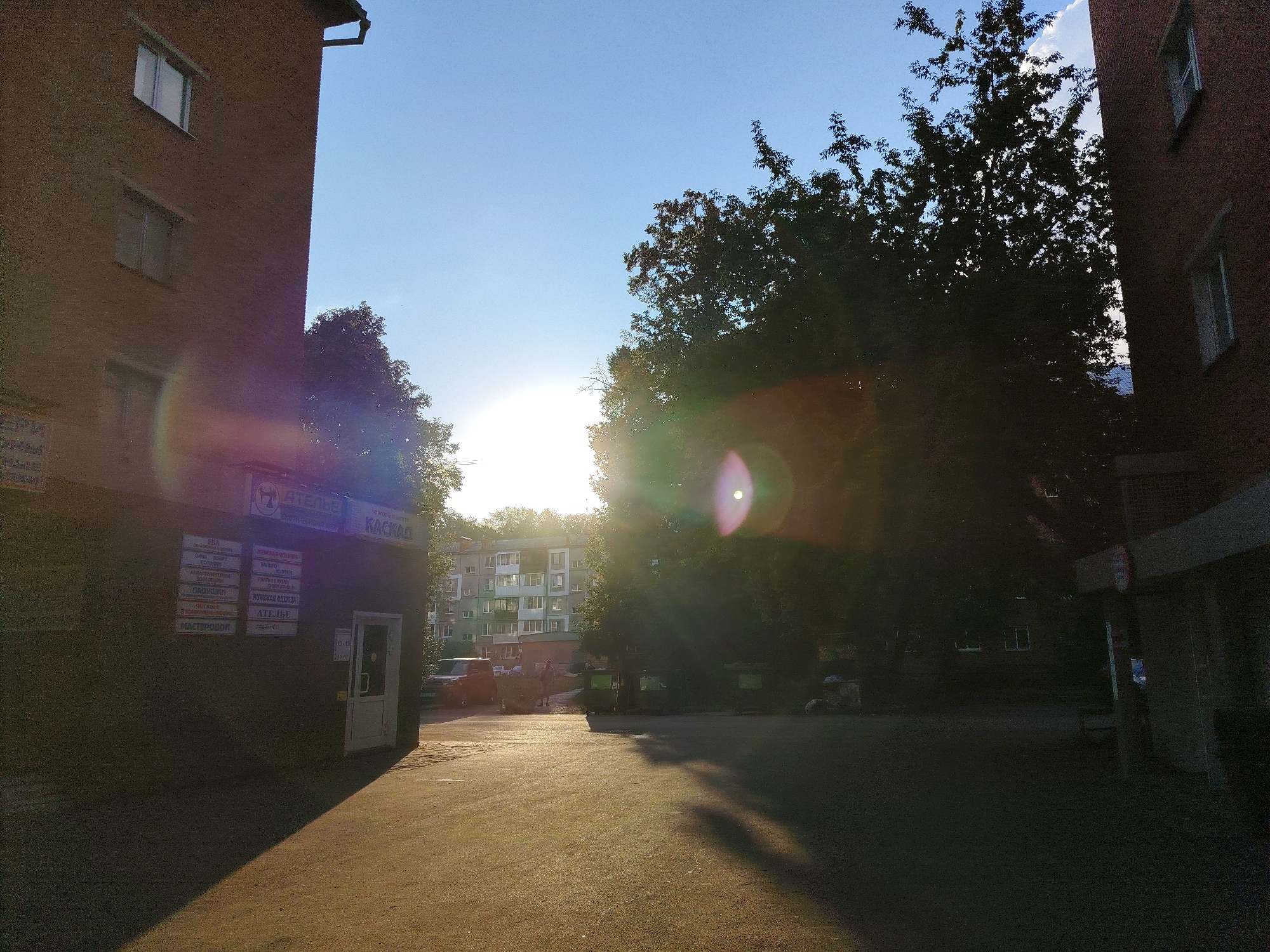 The image depicts an outdoor scene in Kemerovo, Russia, likely in the late afternoon or early evening due to the low angle of the sun. The perspective is from a street or courtyard, looking towards a bright sun that is partially obscured by trees.
In the foreground, a paved surface, possibly asphalt, is illuminated by the setting sun, casting long shadows. To the left, a portion of a brick building is visible, featuring multiple windows. A sign is affixed to this building, indicating services such as "АТЕЛЬЕ" (Atelier/Tailor Shop) and "КАСКАД" (Cascade). Several smaller signs list various services or products. A white door is present in this section of the building.
Dominating the mid-ground and extending into the background are large, dark green trees, their foliage silhouetted against the bright sky. The sun's glare creates lens flare effects, appearing as bright circular or oval shapes with halos.
In the background, a multi-story apartment building with numerous windows is visible. A few vehicles are parked or driving in the vicinity of the apartment building and near the atelier. There are also several large green trash bins visible. A person is walking in the distance near the apartment building.
The sky is a clear, pale blue, with a few wisps of white clouds visible towards the top right. The overall impression is an urban environment bathed in the warm light of a setting sun.