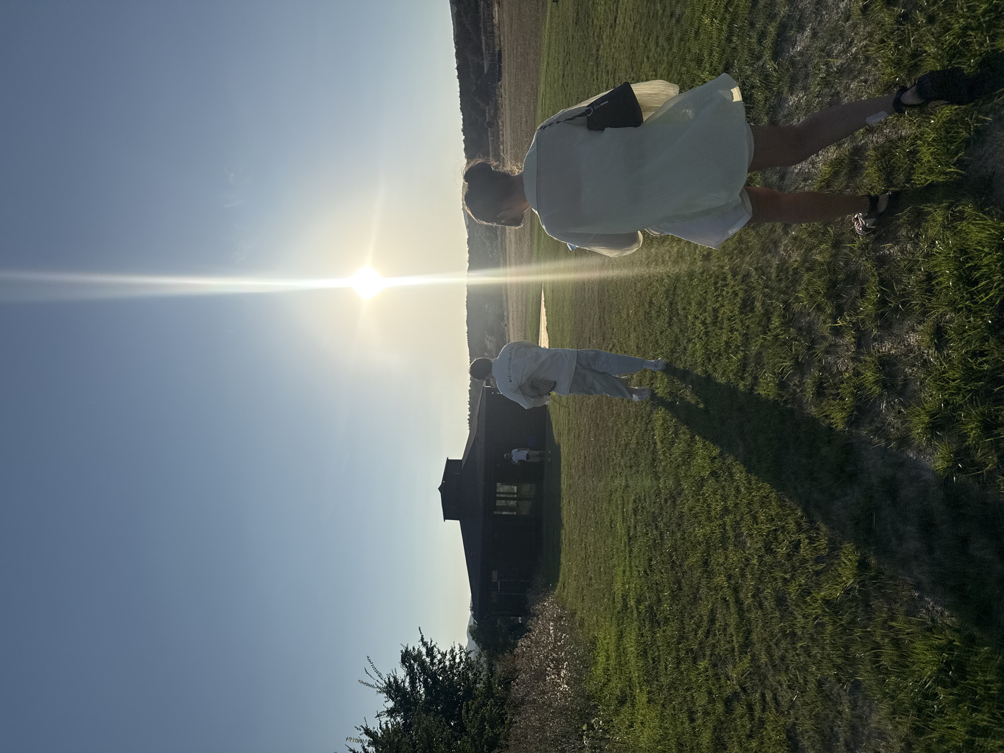 In Orlinovsky District, Ukraine, the image captures a scene on a grassy field under a bright, sunny sky. The sun is low on the horizon, casting a long shadow of a person walking towards the viewer's right. This individual is wearing a light-colored, possibly white, shirt and dark shoes. Further into the field, a dark-colored building with a sloped roof and windows is visible. Another person, also wearing light-colored clothing, is walking towards the building. The environment appears to be rural, with rolling hills and vegetation in the background. The overall lighting suggests it is either sunrise or sunset.