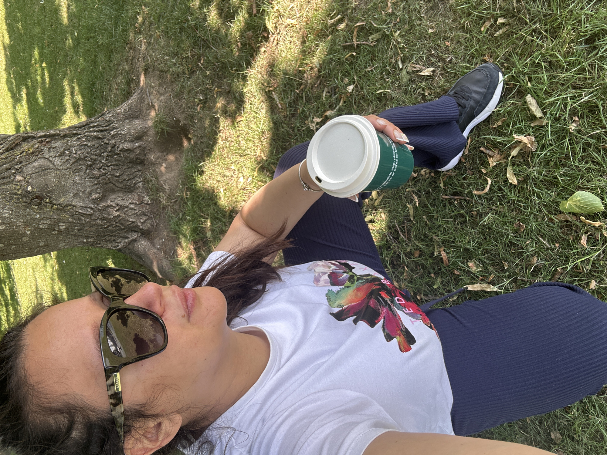 A woman with dark hair, wearing large sunglasses and a white t-shirt with a floral print and navy ribbed pants, sits on the grass next to a large tree. She holds a green reusable coffee cup in her right hand, which is adorned with a silver bracelet. The background consists of a grassy area with scattered leaves and dappled sunlight filtering through the tree canopy. The scene suggests an outdoor leisure activity in a park or natural setting.