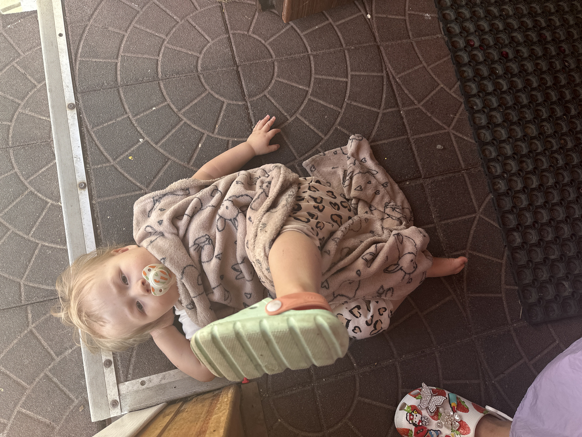 A young child lies on a patterned tile floor, covered by a beige blanket decorated with various cartoon characters and leopard print. The child has blonde hair and a pacifier in their mouth. They are wearing a light green sandal on one foot. The background features a textured rubber mat and part of a doorway. The child is near a threshold with a metal plate. Another pair of adult sandals are nearby with decorative charms on them. The location could be a domestic porch or patio in Nizhny Novgorod.