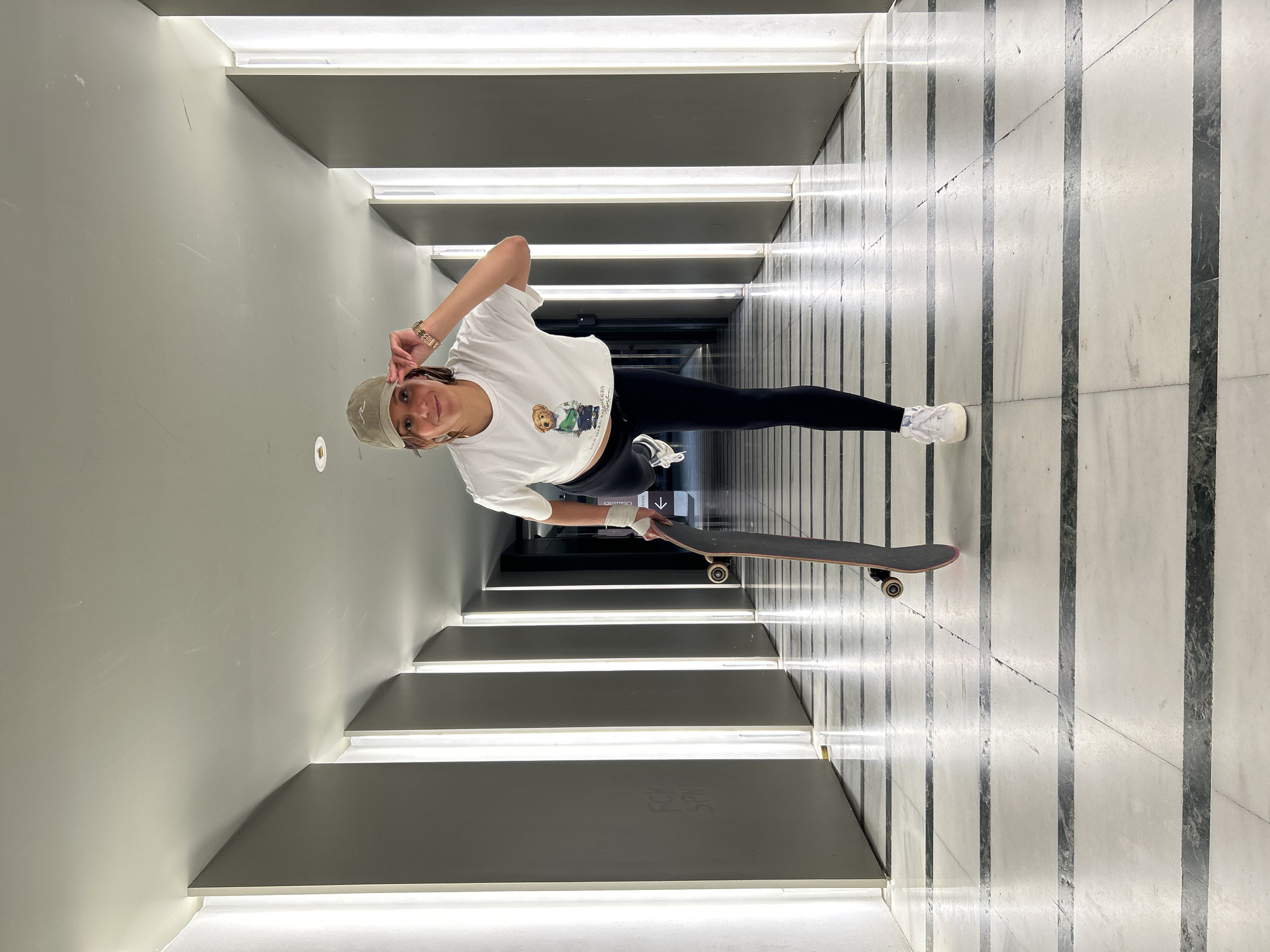 A person wearing a white t-shirt with a cartoon bear on it, black leggings, a khaki baseball cap, and white sneakers is standing in a hallway holding a skateboard. The hallway has a light grey painted wall on the left and a white and grey marbled wall on the right. There are horizontally placed lights installed into the wall on the left-hand side. The hallway appears to lead outside to a bright, light-filled area. The person is smiling and making a hand gesture near their head. Based on the architecture and interior design, the location might be in Barcelona, Spain.