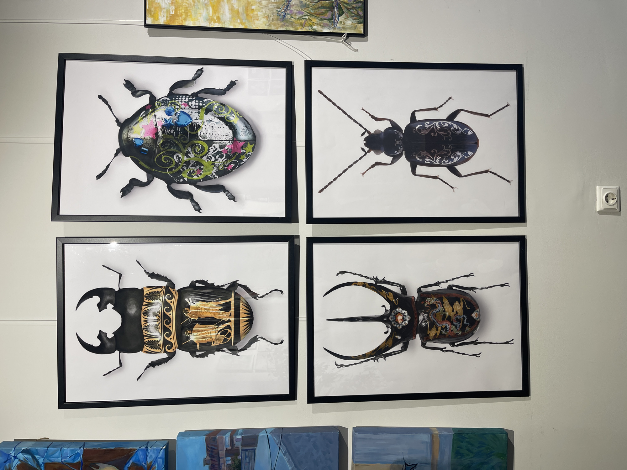 The image shows a display of four framed artworks of beetles hanging on a light-colored wall, possibly in a gallery or exhibition space. The beetles are presented in a detailed and decorative style. The artworks are arranged in a square formation, and some other paintings are visible at the bottom of the frame. A white electrical outlet is visible on the right wall. This image was taken in Sofia, Bulgaria.