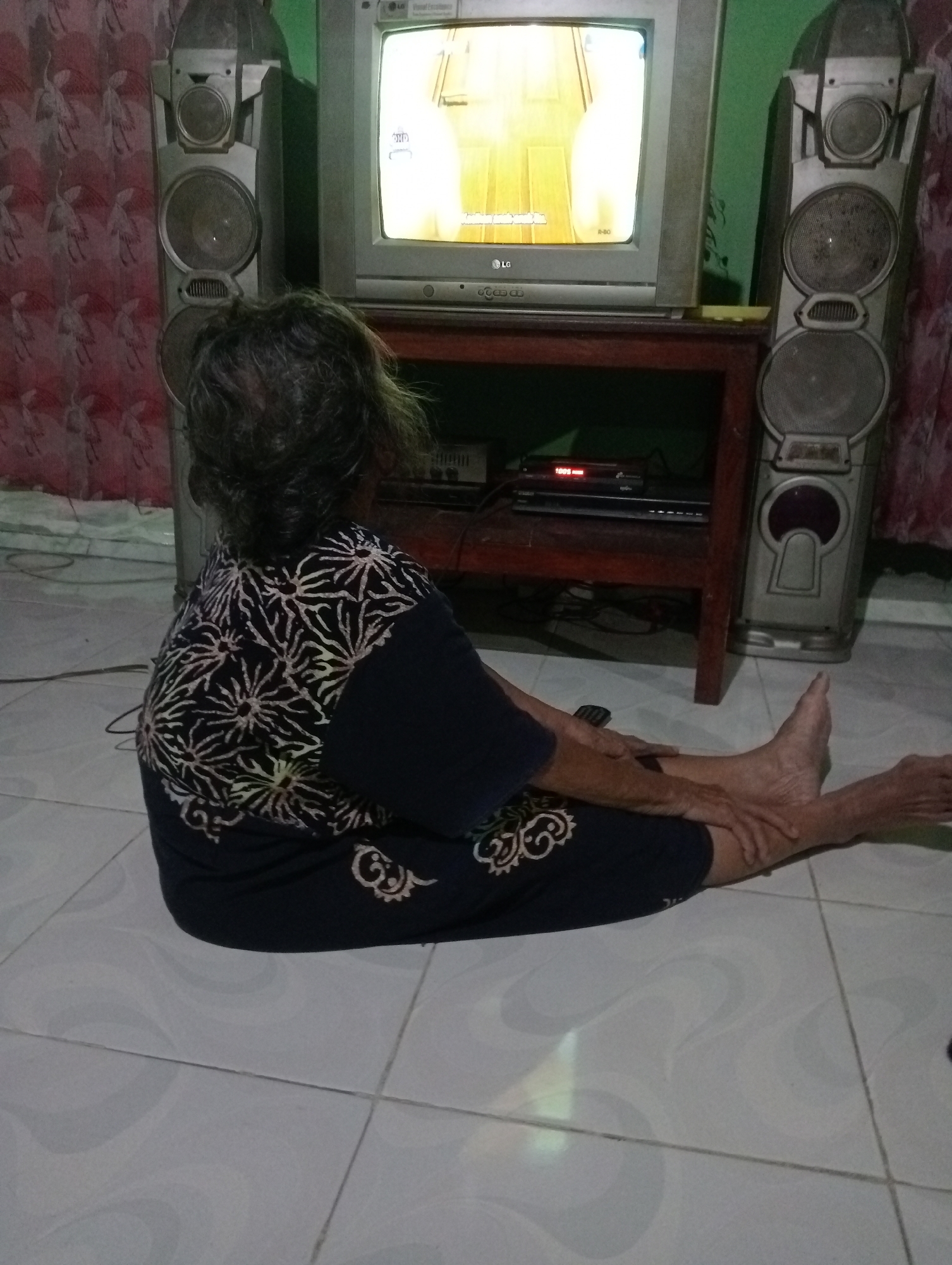 In a dimly lit room with tiled floors, an elderly person with gray hair is seated on the floor, facing away from the camera. They are wearing a dark blue garment with a white floral pattern. Their legs are extended, and one foot is visible, appearing bare. The person is looking at a television screen, which is displaying a bright image. Beside the television are two large, silver speakers, one on each side. A wooden stand holds the television and a DVD player is visible beneath it. The room is decorated with a patterned curtain on the right side. The scene is likely in Kota Negara Timur, Indonesia, given the context of the location.
