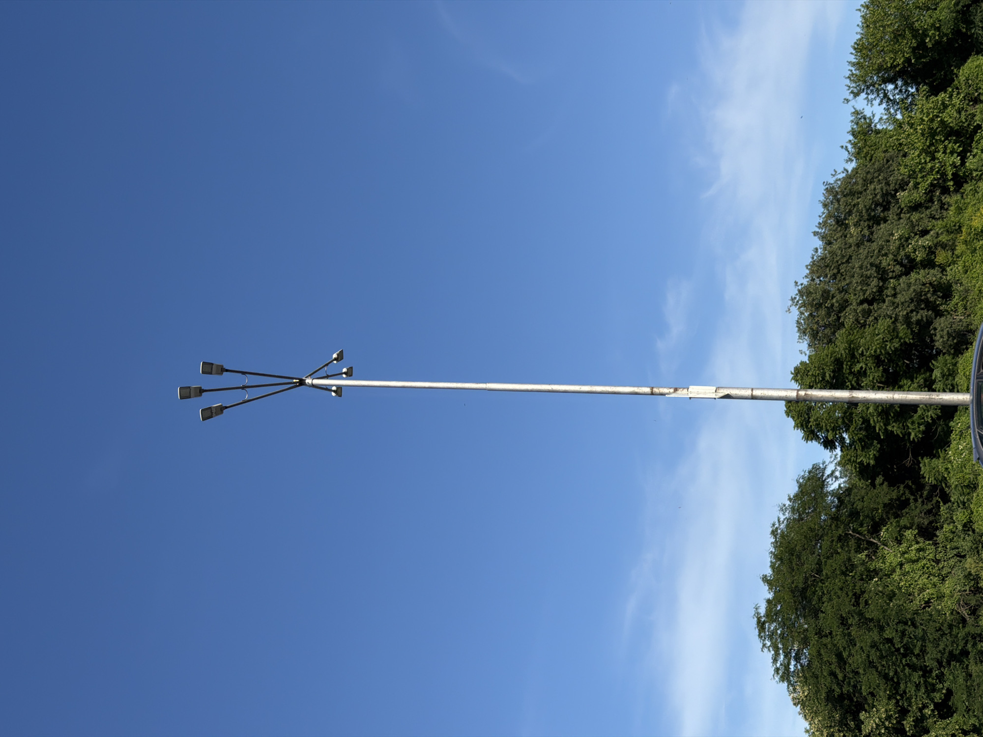 This media file is a photograph taken in Burgas, Bulgaria. The image primarily features a bright blue sky with some wispy white clouds. A tall, slender pole, likely a streetlamp, extends vertically through the frame, with multiple light fixtures clustered at its top. The light fixtures are angled outwards, suggesting they are designed to illuminate a wide area. To the right of the pole, a dense canopy of lush green trees fills the frame, indicating a natural or park-like setting. The perspective is from below, looking upwards towards the sky and the top of the pole. The sunlight suggests it is daytime, and the clear sky implies fair weather. There is no discernible activity or event taking place; the scene is static and focuses on the composition of the sky, the pole, and the trees. No text is visible in the image. The overall impression is one of an outdoor scene on a clear day, with man-made elements like the streetlamp integrated into a natural landscape.