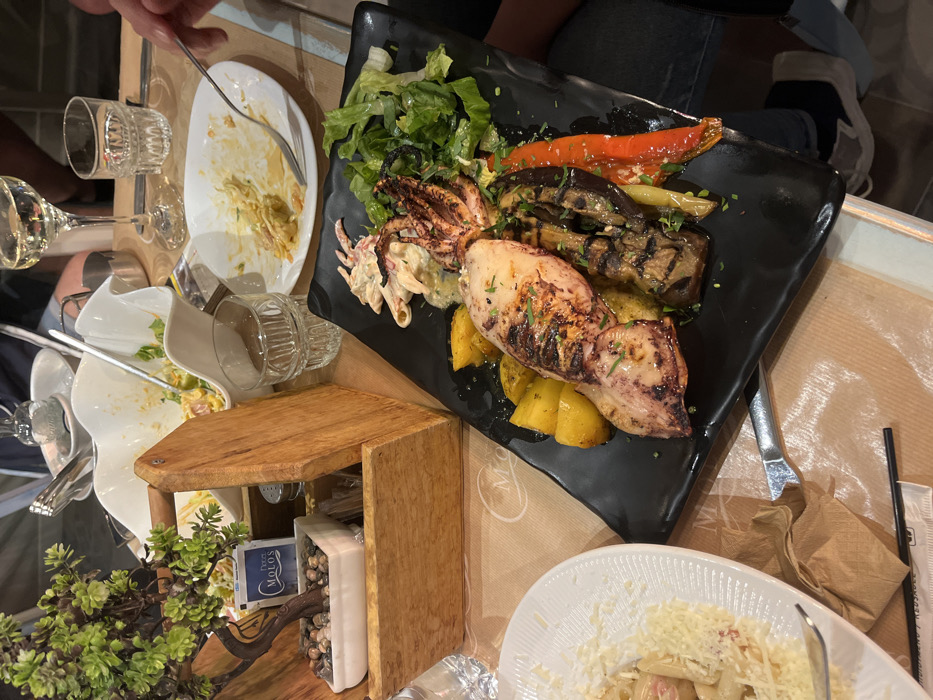 The scene is a table at a restaurant in Limenaria, Greece (latitude: 40.628, longitude: 24.575). A plate of food is in the center of the table, containing grilled squid, eggplant, peppers, and potatoes. There are also plates of food, a glass of wine, and other miscellaneous items on the table. The person in the background is partially visible, wearing jeans. The environment is a casual restaurant setting.