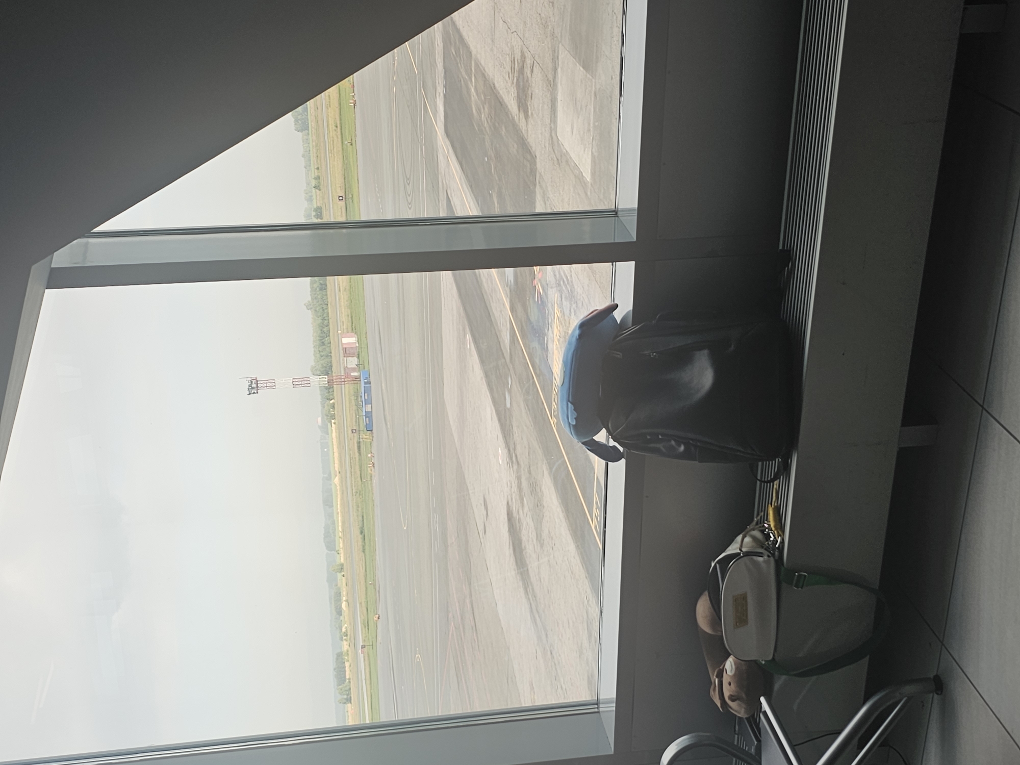 A view from an airport window in Ob, Russia, looks out onto an airfield. A tall red and white radio tower stands in the distance. In the foreground, on a bench, are two backpacks. One is dark and sits upright, while the other, a lighter color, is placed in front of it. A stuffed animal is also visible on the bench. The lighting suggests it is daytime, and the sky is overcast. The scene is typical of an airport waiting area.