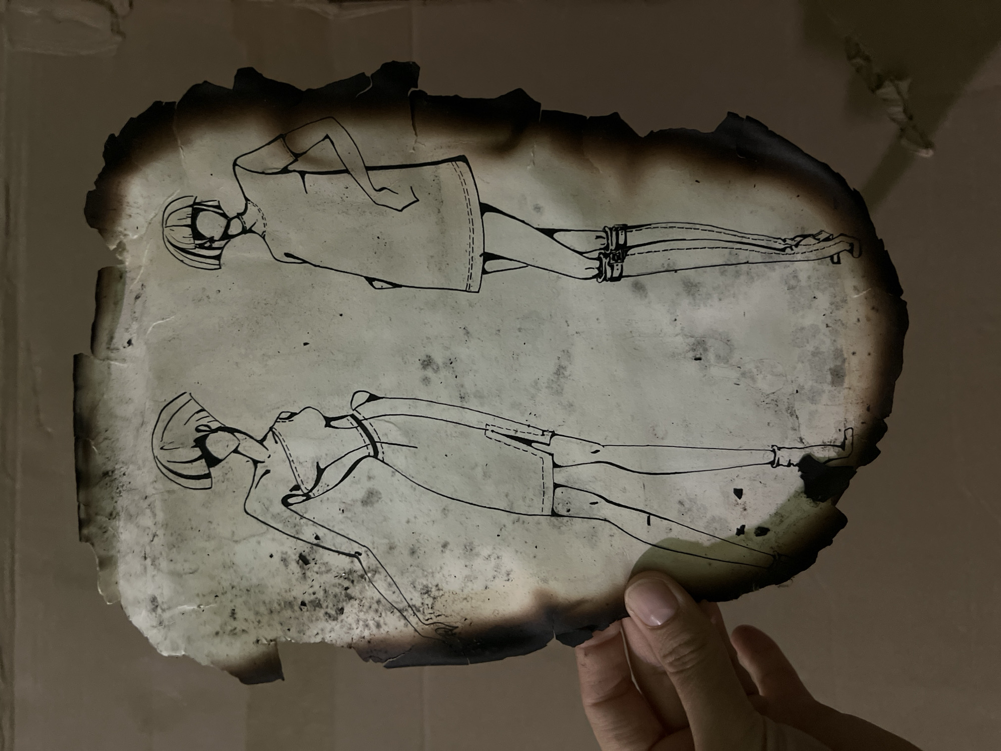 A hand holds a burnt drawing of two fashion figures against a plain brown background. The drawing, rendered in black ink, depicts two stylized female figures with short, geometric haircuts and faceless features. The figure on the left wears a strapless dress with a high slit and what appears to be a belt or sash. The figure on the right is in a short, A-line dress with visible stitching details and knee-high boots. The paper is significantly charred and damaged around the edges, with some smudges and discoloration present on its surface. The background appears to be a wall or large surface, illuminated by ambient light, creating shadows that fall behind the drawing. No specific location in Moscow, Russia is identifiable.