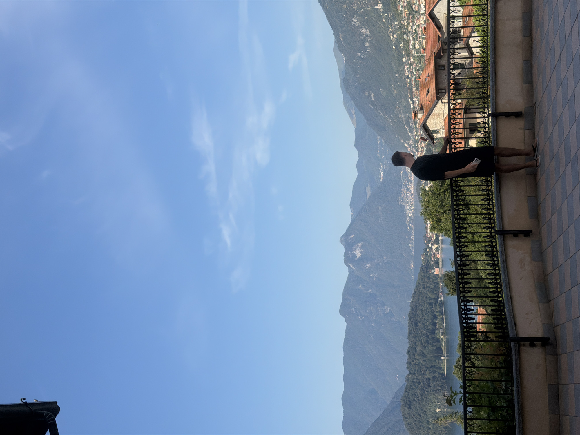 A man stands on a tiled balcony overlooking a town nestled between a lake and mountains. The man is wearing a black t-shirt and shorts, holding a phone in his right hand. The balcony has a decorative metal railing and a stone wall. The town below features terracotta-roofed buildings and is surrounded by lush green trees and hills. In the distance, a large lake is visible, with a chain of mountains forming the backdrop. The sky is a clear blue with scattered wispy clouds. The scene is likely Tremezzina, Italy, given the characteristic architecture and landscape.