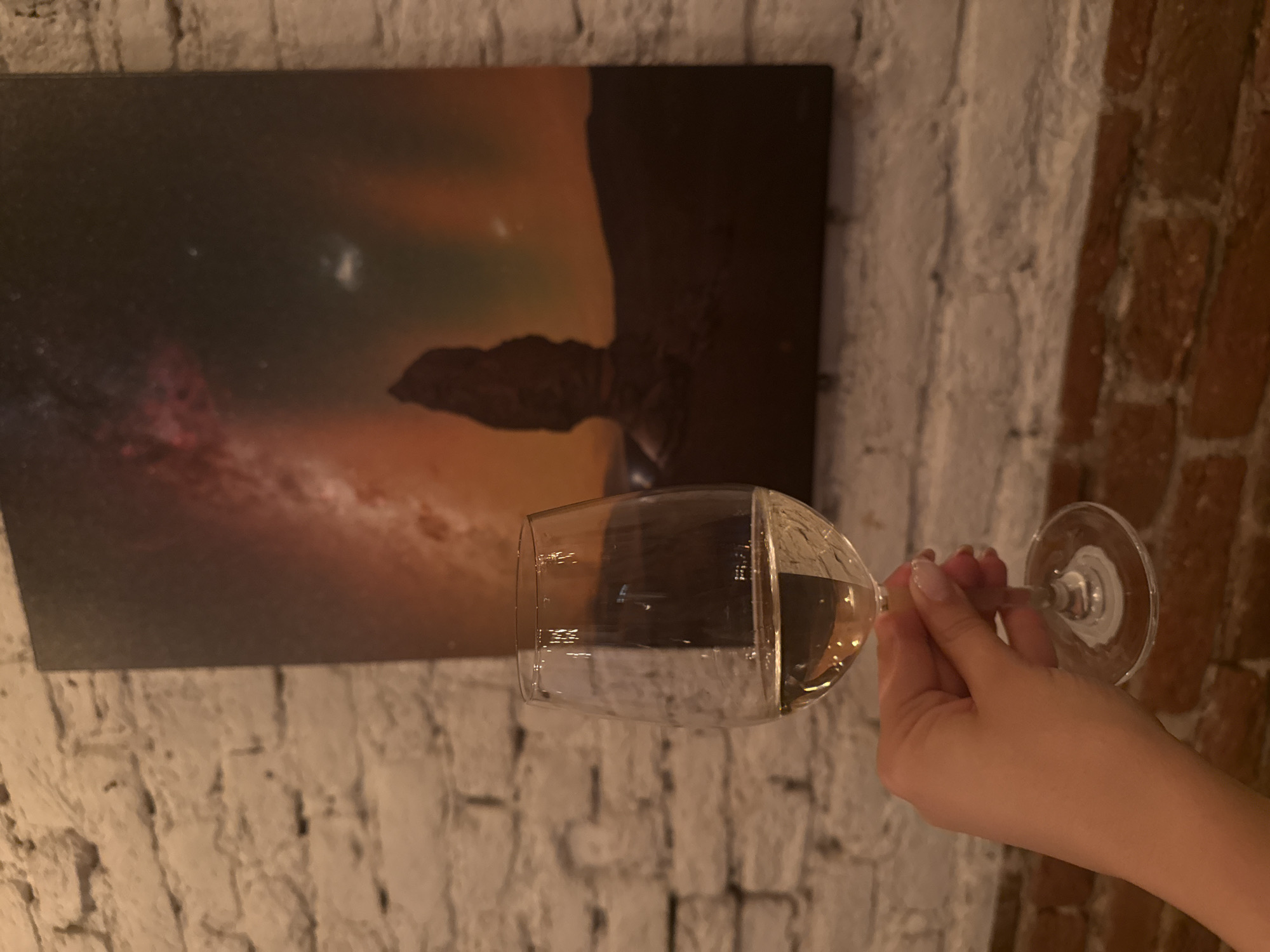 A person is holding a glass of white wine in front of a textured white brick wall, with a painting featuring a celestial scene hanging on the wall. The right side of the wall is a red brick wall. The location could be in Sofia, Bulgaria. The hand of the person holding the glass is visible. The wine is transparent and fills about a third of the glass.