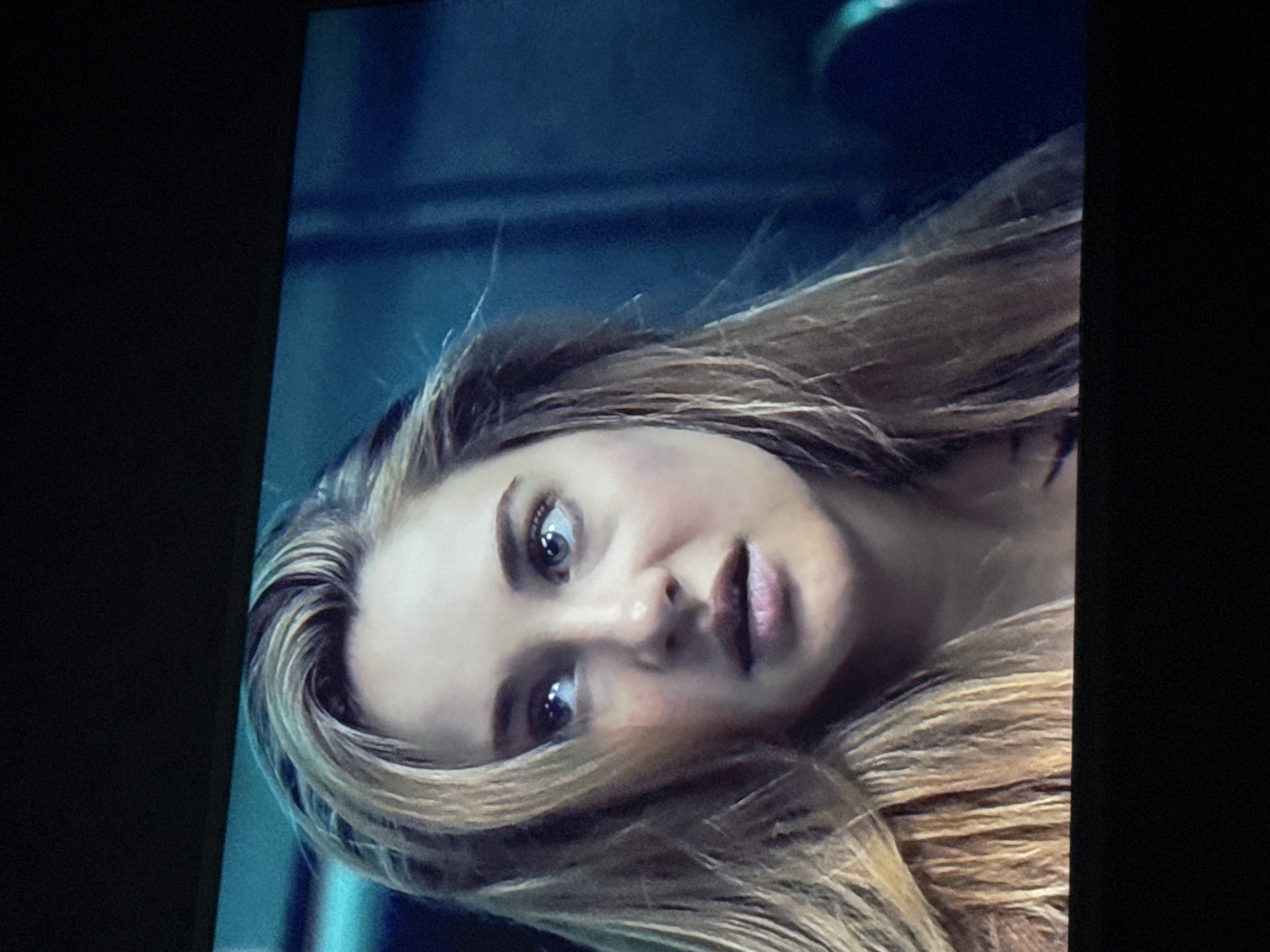 A close-up shot of a woman's face, possibly on a television screen, is oriented at an angle. The woman has light skin, light brown hair with blonde highlights, and a slightly open mouth. The background appears to be a blurred, dark blue color. There is no identifiable location in Mytishchi, Russia, or any city visible in the image.