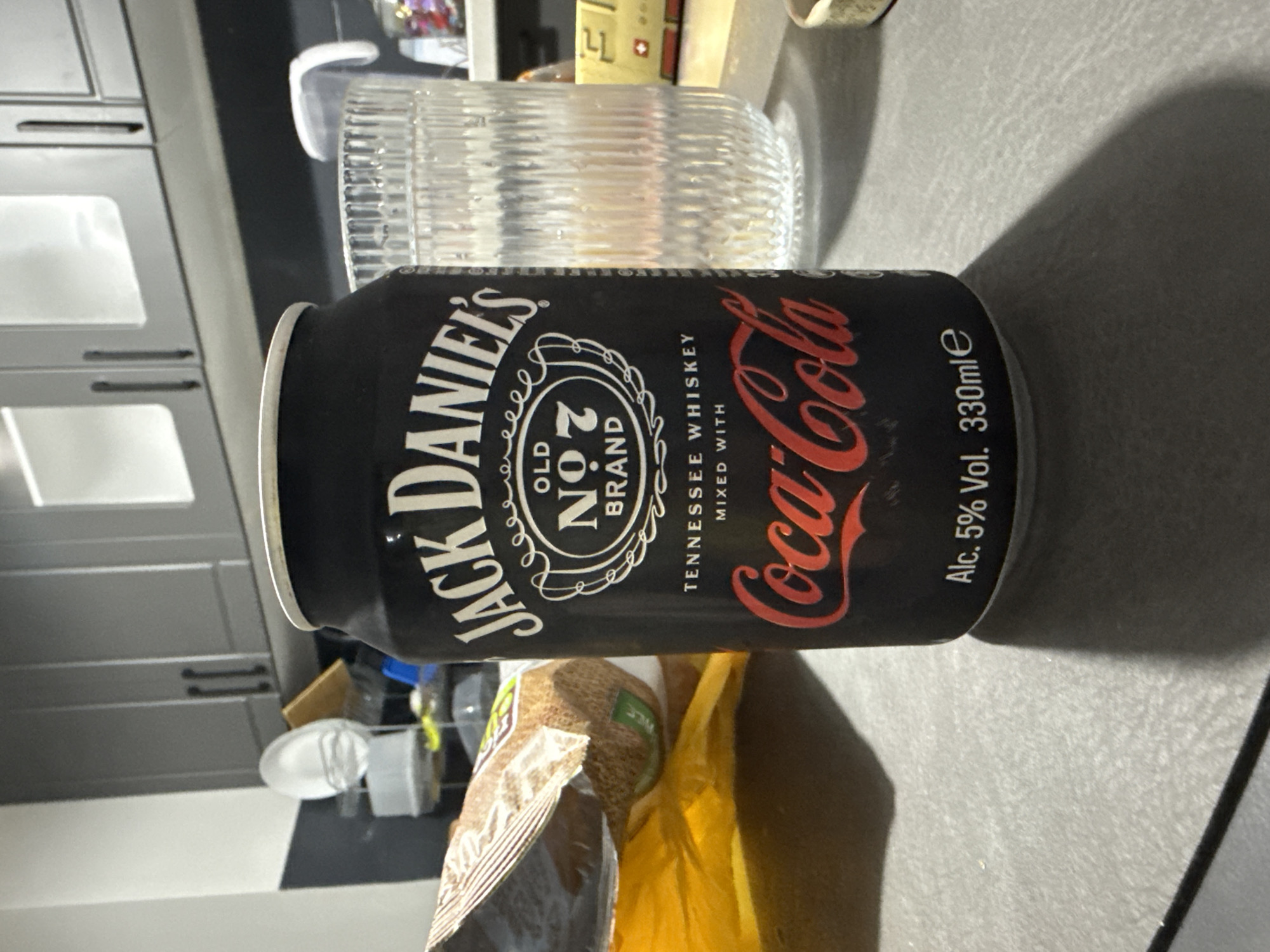 An indoor, close-up shot captures a black can of Jack Daniel's & Coca-Cola ready-to-drink beverage standing upright on a speckled grey countertop. The can features "JACK DANIEL'S OLD NO. 7 BRAND" in prominent white lettering, with "TENNESSEE WHISKEY MIXED WITH" stated below. The distinctive red "Coca-Cola" logo is displayed on the right side of the can. At the bottom, "Alc. 5% Vol. 330mle" is visible, indicating the alcohol content and volume.
Beside the can, to its left, a tall, clear glass with a ribbed design is also on the countertop. In the background, modern grey kitchen cabinets with dark handles suggest a kitchen or bar setting. The lighting appears to be artificial. In the foreground, partially visible, are open packages of what appear to be snacks or food items, one in a brown and white striped bag, and another in a yellow bag. A dark shadow is cast on the countertop to the right of the can. The scene suggests a casual moment, possibly for enjoying a beverage in Stara Zagora, Bulgaria.
