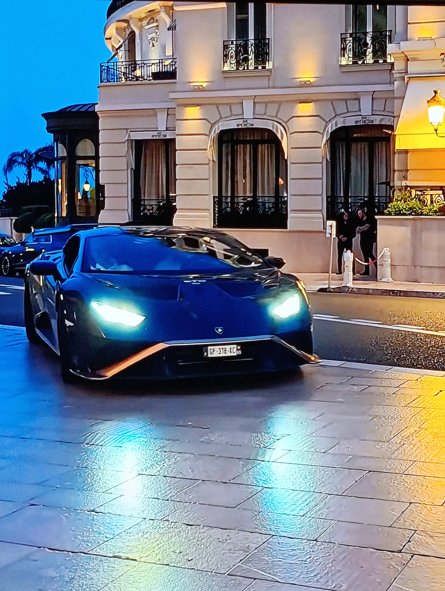 A dark blue Lamborghini with its headlights on is parked on a wet, tiled street in front of a light-colored building. The building has multiple arched windows with dark curtains, and decorative wrought-iron balconies. Two individuals in dark clothing stand near a white bollard and a red-and-white barrier on the sidewalk beside the building. Another vehicle is faintly visible in the background. The license plate of the Lamborghini reads "GP-378-KC". The environment suggests a European city at dusk or early evening.