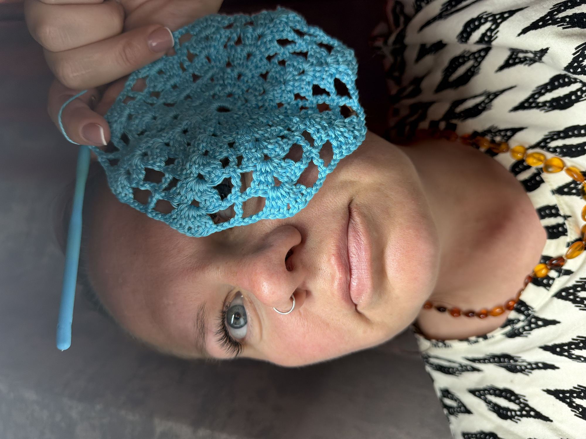 A person with fair skin, blue eyes, and a nose ring is holding a blue crocheted doily over their forehead. The person is wearing a black and white patterned shirt and an amber-colored beaded necklace. A blue crochet hook is also visible. The photograph appears to have been taken indoors. There is no evidence to suggest that the photograph was taken in Chaikovsky, Russia.