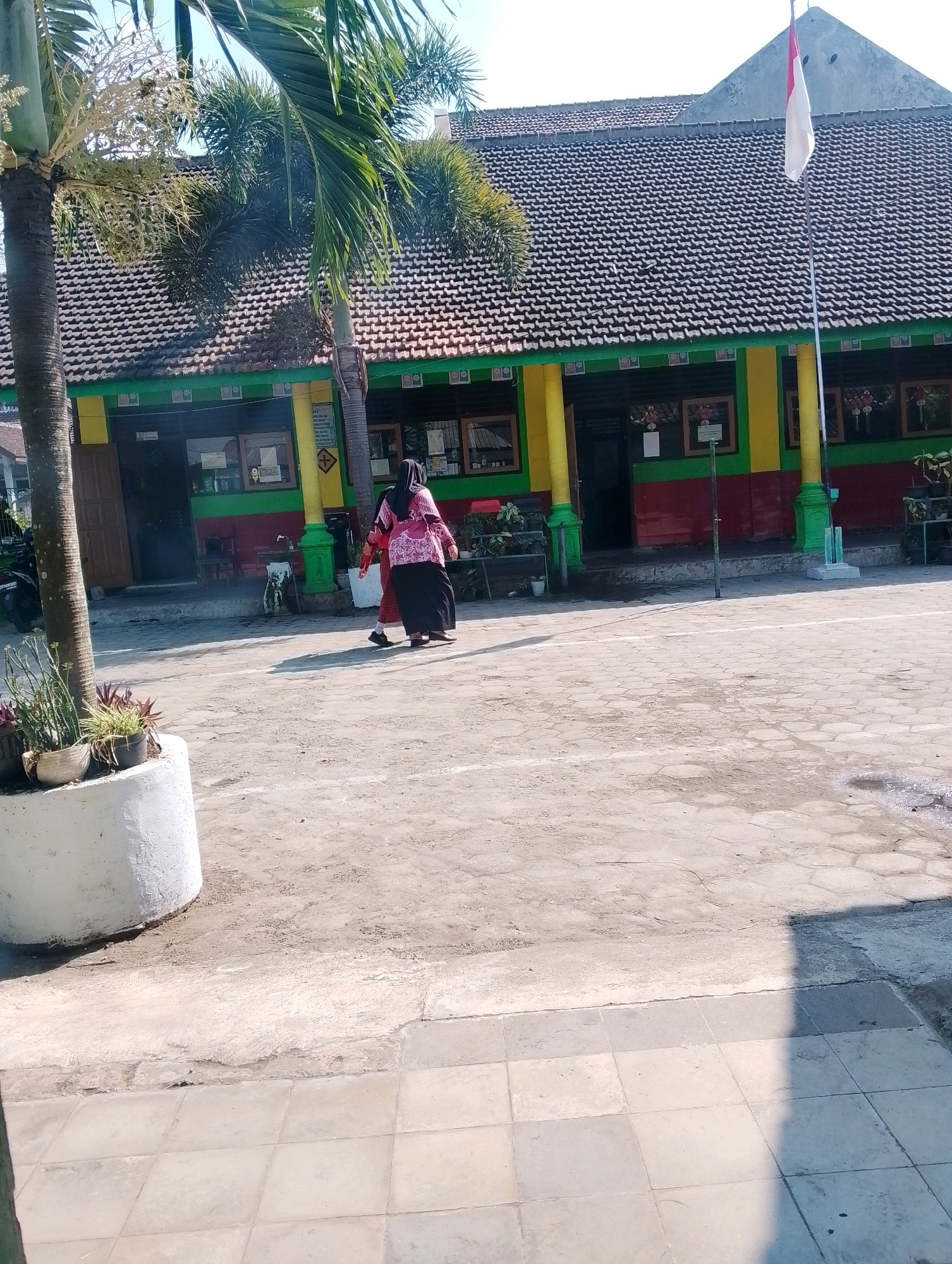 The image depicts the exterior of a school building, likely in Papungan, Indonesia, under a bright, clear sky. Two individuals, presumably students or staff, are walking away from the camera across a paved courtyard. One person is wearing a pink headscarf and a long-sleeved top with a patterned design, paired with a black skirt. The other person is in a red long-sleeved top and a dark skirt.
The building's facade is painted in vibrant colors: red on the lower section, a green band above it, and yellow pillars supporting an overhang. The roof is covered with terracotta tiles. A flagpole with the Indonesian flag, red and white, is prominently displayed on the right side of the building. In the foreground, a large white planter contains various green plants. The courtyard surface is a mix of hexagonal paving stones and concrete. To the left, part of a palm tree is visible, with its fronds extending into the upper left corner of the frame. A motorcycle is parked to the far left, partially obscured by the palm tree. The overall impression is that of a typical Indonesian school environment during daylight hours.