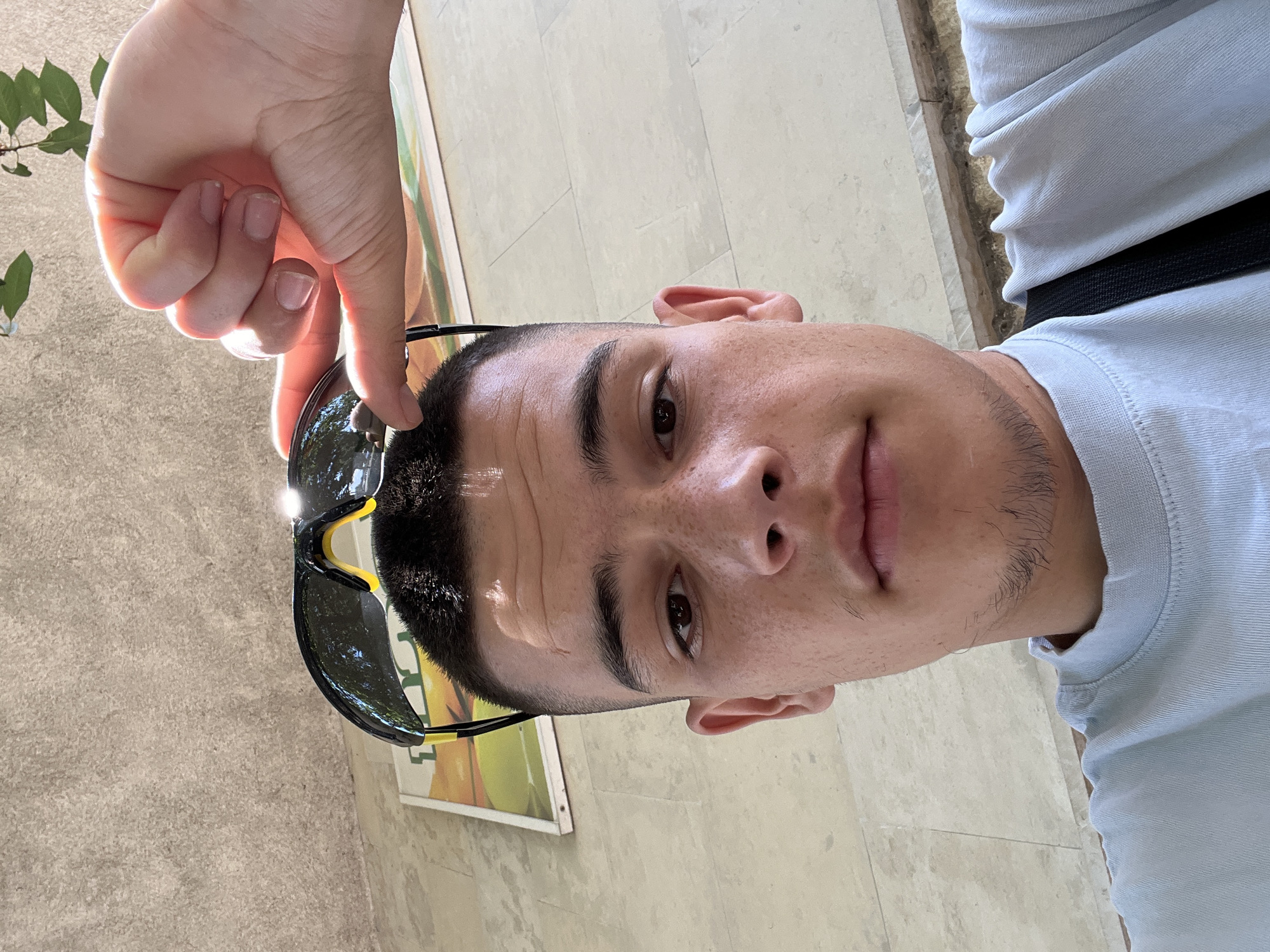 In a close-up, outdoor, waist-up selfie, a young man with a short buzz cut and freckled skin holds a pair of sunglasses with his right hand on top of his head. He is looking directly at the camera with a neutral expression. The background consists of a textured stone wall with a framed poster partially visible behind him. A hint of green foliage is seen in the upper right corner. The overall lighting suggests it is a bright, sunny day. The subject is wearing a light blue t-shirt.