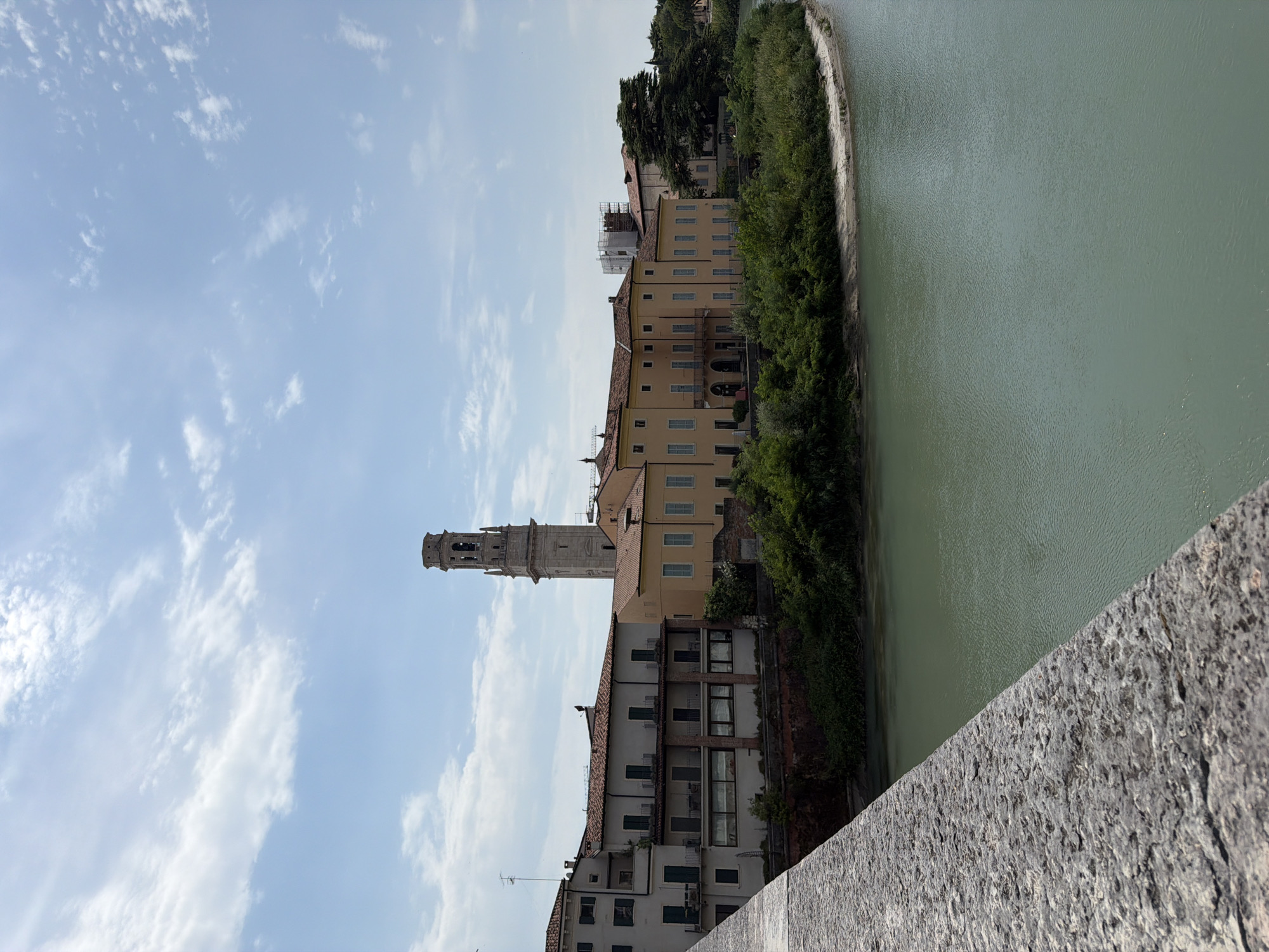 An outdoor, eye-level, vertical medium shot captures a view of the Adige River in Verona, Italy. The river occupies the majority of the lower half of the frame, its waters a muted green with subtle ripples indicating a gentle current. In the foreground, a stone embankment runs diagonally from the lower left to the mid-left, its rough texture and grey hue contrasting with the smoother surfaces of the buildings and water.
Across the river, a line of historic buildings runs parallel to the water's edge. These structures, with their terracotta tiled roofs and varying pastel facades—ranging from pale yellow to off-white—appear to be several stories tall. Balconies with dark railings punctuate the facades. To the left, a building features a more muted color scheme and visible antennas on its roof. To the right, a tall, slender bell tower with arched openings and decorative stonework rises above the other buildings, characteristic of Italian architecture. Lush green trees and vegetation line the riverbank between the buildings and the water, with a sliver of sandy bank visible in the far right. The sky above is a pale blue, dotted with wispy white clouds. The scene is bathed in natural daylight, suggesting it was captured on a clear or partly cloudy day. No people are visible in the frame.