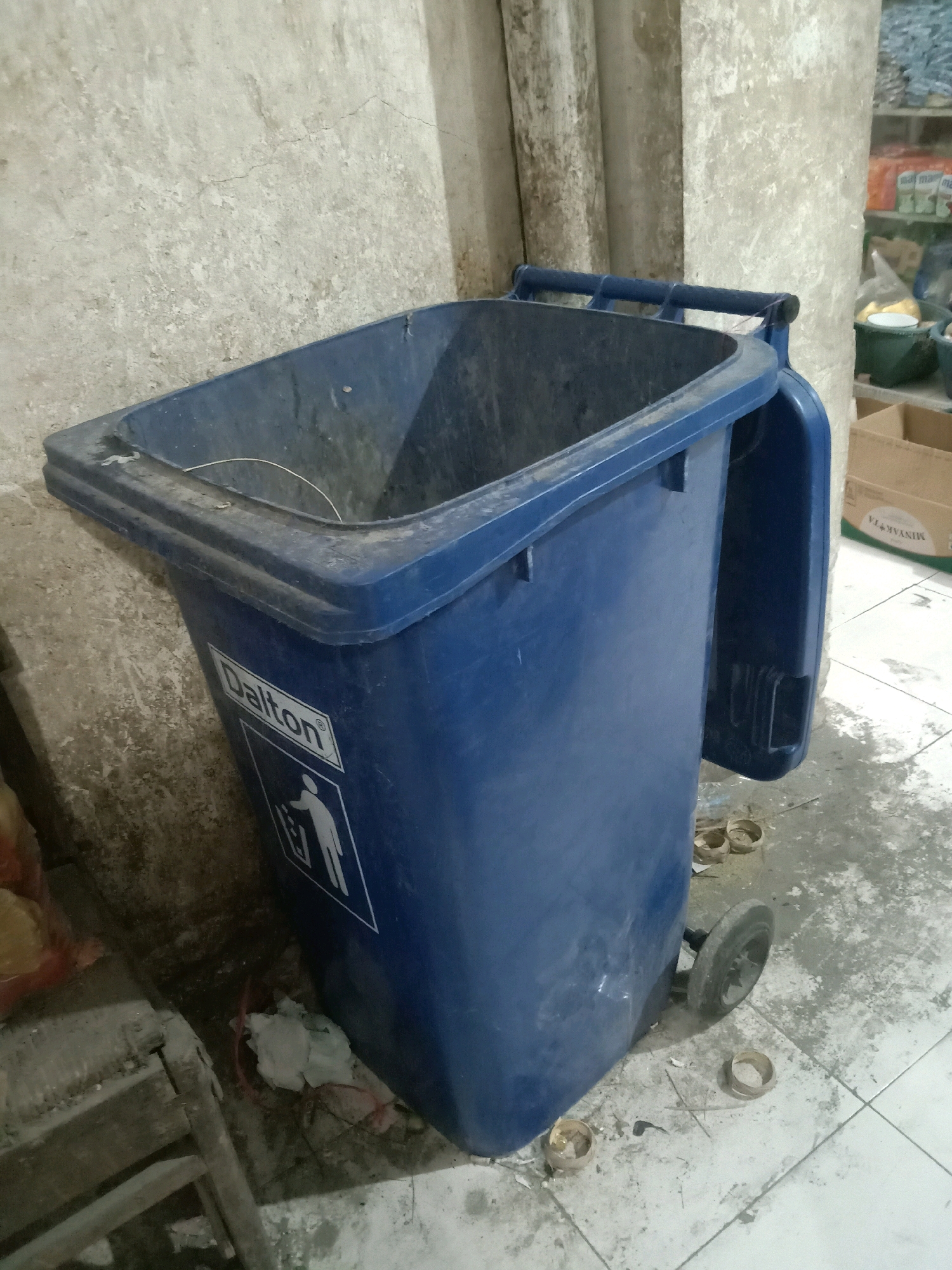 A blue Dalton brand trash can with wheels is situated in an outdoor area. The trash can is open and appears empty, with a wire hanger inside. It is positioned on a tiled floor next to a rough, possibly concrete, wall. Debris, including scattered paper and what looks like dried leaves, is present on the ground around the trash can. In the background, shelves containing various items are visible, along with cardboard boxes. The scene suggests an outdoor storage or utility area, possibly in a residential or commercial setting within the city of Sugihwaras, Indonesia.