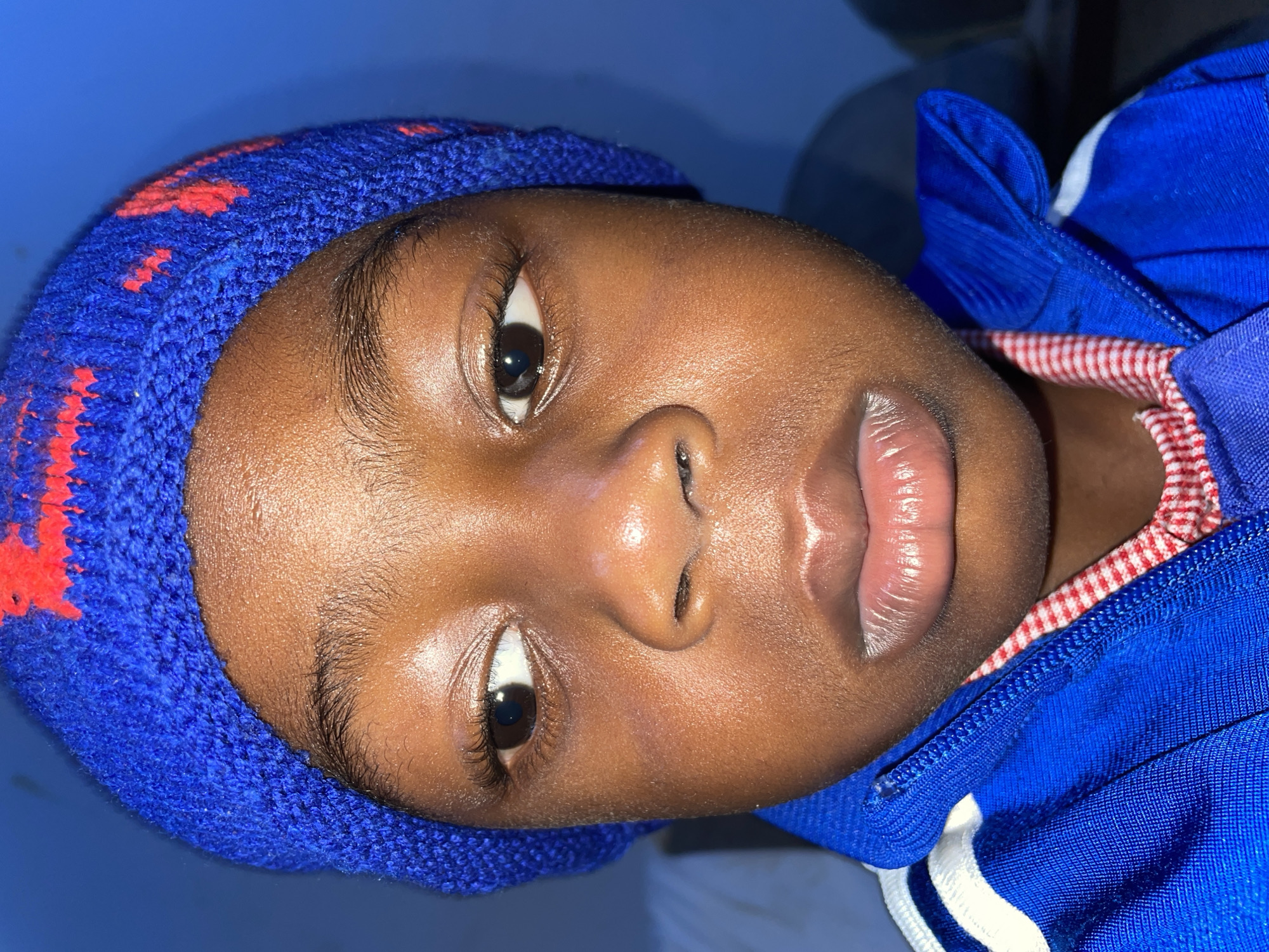 A close-up portrait depicts a young person with dark skin, wearing a blue knitted hat with red accents. The individual also wears a blue and white jacket over a red and white checkered shirt. The photo's background consists of a blue surface. There's no explicit information in the image to confirm its location as Jos, Nigeria.