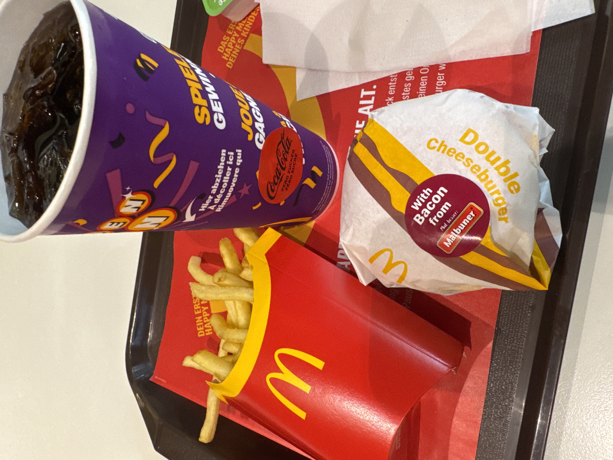 The image displays a McDonald's meal served on a black tray. The meal includes a large purple cup filled with a dark liquid, likely Coca-Cola Zero, with German and French text on the cup. There is also a box of McDonald's fries. Additionally, there is a "Double Cheeseburger with Bacon from Malbuner". The food is sitting on a red paper liner with German text and McDonald's logos. Based on the text "DEIN ERSTE HAPPY MEAL DEINES KINDES" and the branding in German, the location is likely a McDonald's in a German-speaking region of Switzerland. Due to the presence of Malbuner bacon (from Switzerland), it is more specifically likely to be a McDonald's in Switzerland, and most likely in the city of Spreitenbach, Switzerland.