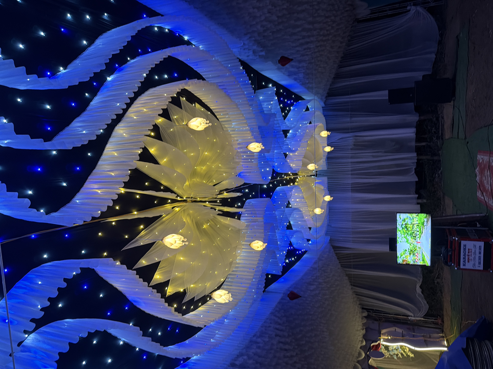 The image captures a vibrant and elaborately decorated ceiling or canopy, likely part of an event venue or temporary structure. The primary decorative elements are large, stylized floral or star-shaped light fixtures, made of translucent white or yellow material and illuminated with warm yellow light. These are arranged centrally, with several individual illuminated fixtures hanging down.
Flowing white and light blue fabric drapes create a wave-like or cloud-like effect across the ceiling, interspersed with numerous small LED string lights in both blue and warm white, giving a starry night sky impression against a dark background.
Towards the right side of the frame, outside the main decorative area, a flat-screen television is visible, displaying a bright outdoor scene. Below the TV are black audio speakers, and next to them, a stack of equipment cases. One case clearly displays the text "KARAOKE," along with other smaller, less legible text.
The scene suggests a festive event, possibly a wedding reception, party, or public celebration, taking place at night or in a low-light indoor setting. The extensive lighting and decorations create a bright and celebratory ambiance. The presence of karaoke equipment indicates entertainment is a key part of the event. The location context is Bình An, Vietnam.
