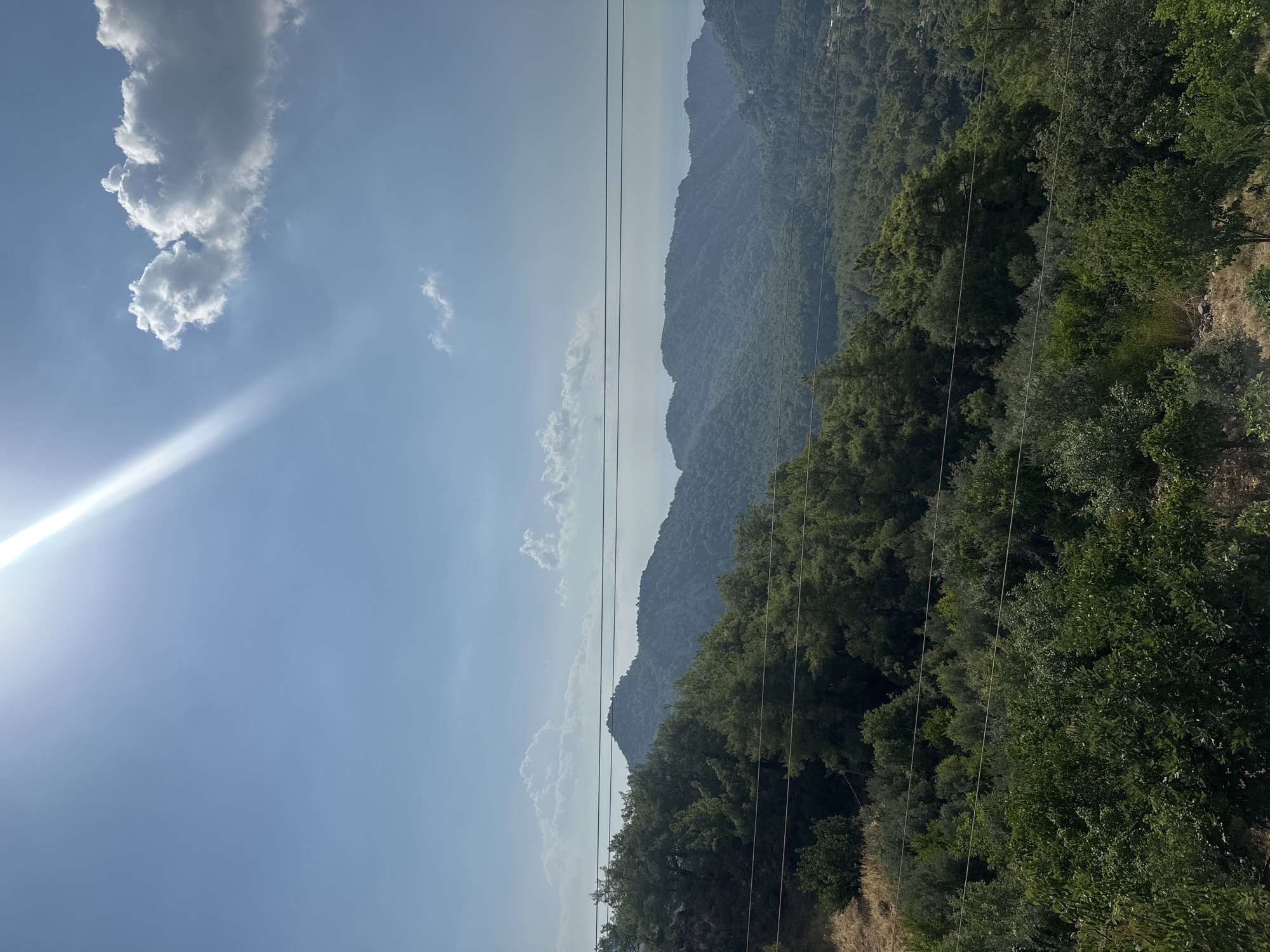 An overhead, wide shot captures a view of mountains and a body of water in Kumluca, Turkey. The sun beams down from the top left corner of the frame, casting a bright ray across the clear blue sky, which is dotted with a few white clouds. The mountains are covered in lush green trees, with the peaks receding into the distance and fading into a haze. The water is visible as a sliver on the horizon, suggesting the mountains are near the coast. Three horizontal power lines cut across the middle of the frame, obscuring the view of the mountains. The foreground consists of more trees and shrubs, with dry, brown grass on the left. The scene is peaceful and natural, with no people or animals visible.