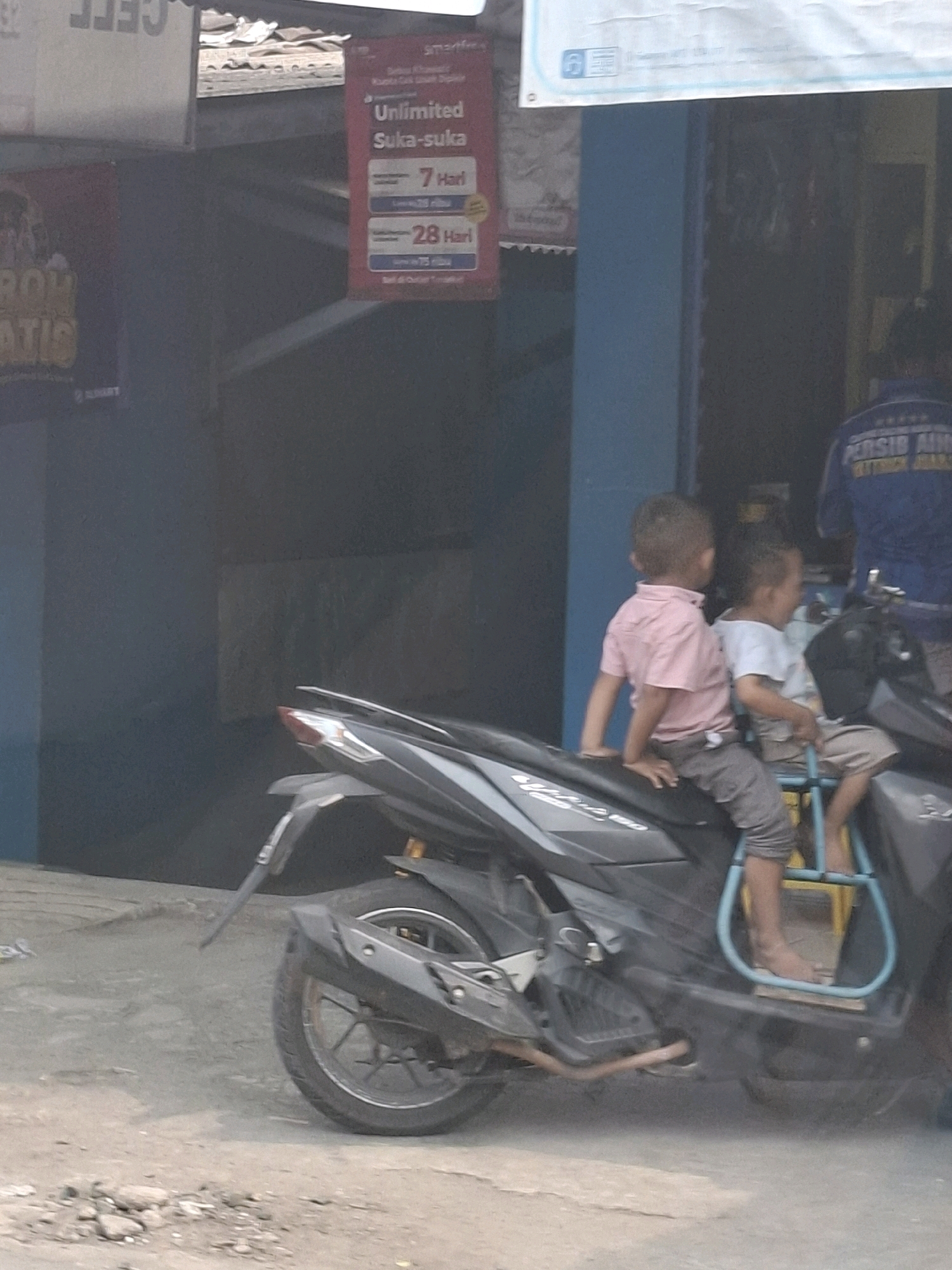 The image captures a scene on a street in Sukabumi, Indonesia. Two young children are seated on a parked black motorcycle. The child in the front wears a light pink collared shirt and grey pants, with their hands resting on the motorcycle's seat. The child behind them wears a light blue t-shirt and beige shorts, positioned in a blue child seat attached to the back of the motorcycle. The motorcycle is parked on a concrete surface, with debris and small stones scattered in the foreground. In the background, a blue building facade is visible, with a red sign hanging from above displaying text related to a mobile network provider offering "Unlimited Suka-suka" with options for "7 Hari" and "28 Hari." Another partially obscured sign with yellow text is also visible. The overall lighting suggests it is daytime.