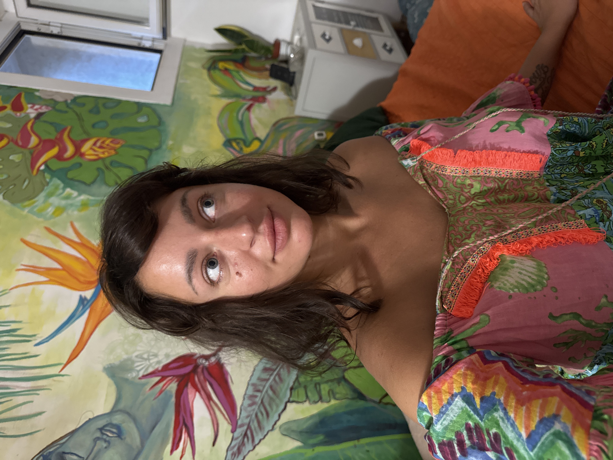 This is a portrait photograph taken indoors, likely in a bedroom, with a vibrant mural as the backdrop. The mural depicts tropical foliage and flowers, including Strelitzia (bird of paradise) flowers, in bold colors like green, orange, red, and blue. A partially opened window with a view of a pale blue sky is visible in the upper left corner. To the right, an orange pillow and a white cabinet with metallic accents can be seen.
The subject is a young woman with dark, shoulder-length hair, lying down and looking directly at the camera. Her expression is neutral, and she appears relaxed. She is wearing a colorful, patterned dress with pink, green, blue, and orange elements, adorned with orange fringe along the neckline and hem. Her right arm, visible towards the top right of the frame, has a tattoo.
The lighting suggests it might be daytime, given the view through the window, but the indoor setting creates a warm, intimate atmosphere. The overall impression is one of a casual, personal moment captured in a colorful and artistically decorated space. There is no visible text in the image. The location context provided is San Benedetto del Tronto, Italy.
