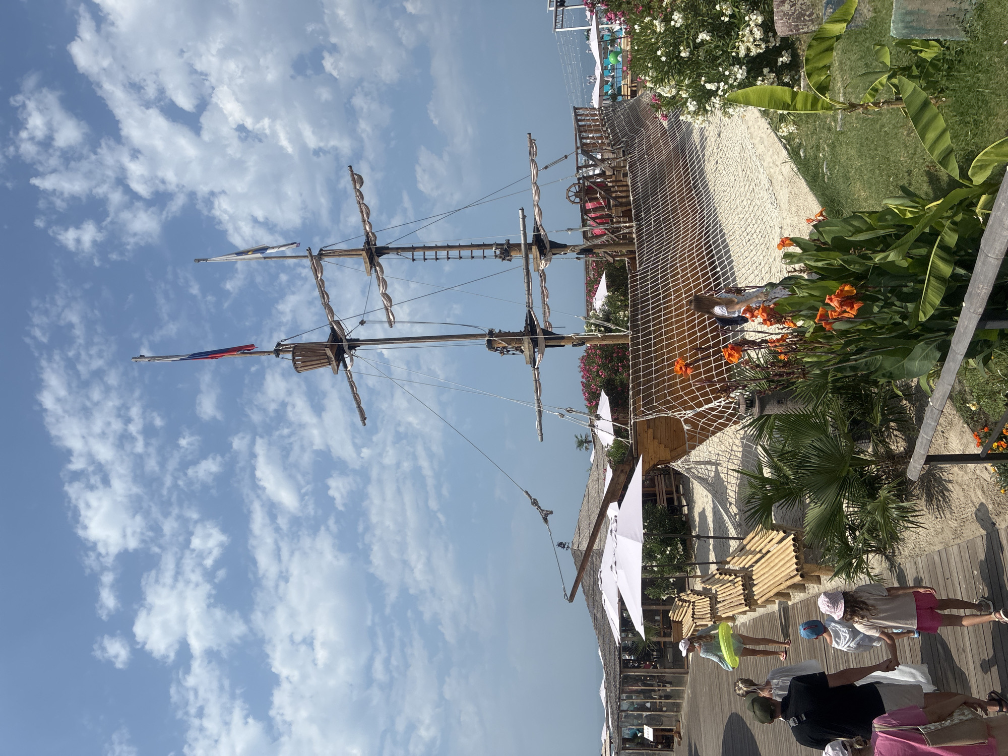 The image depicts a beachside scene in Sirius, Russia, dominated by a large, wooden replica of a pirate ship. The ship, with its masts, rigging, and sails furled, is positioned on a sandy area surrounded by a net enclosure. In the foreground, a wooden boardwalk runs parallel to the beach, with several people walking along it.
On the boardwalk, a group of people are visible. A man in a dark t-shirt and shorts walks ahead, holding a strap of a bag over his shoulder. Behind him, a woman in a white dress and a child in pink shorts and a light-colored t-shirt walk hand-in-hand. Further back, a woman in a light blue top and a white skirt holds a green inflatable ring. Several other individuals, including children, are also present, some walking and some standing.
Adjacent to the boardwalk, there are benches made of bamboo-like material arranged in a curved fashion. White umbrellas are set up over some of these benches, providing shade. Lush green vegetation, including palm-like plants and flowering bushes with orange and white blooms, surrounds the scene.
The background shows the sea and the sky. The sky is blue with scattered white clouds. The overall atmosphere is that of a sunny day at a recreational area.