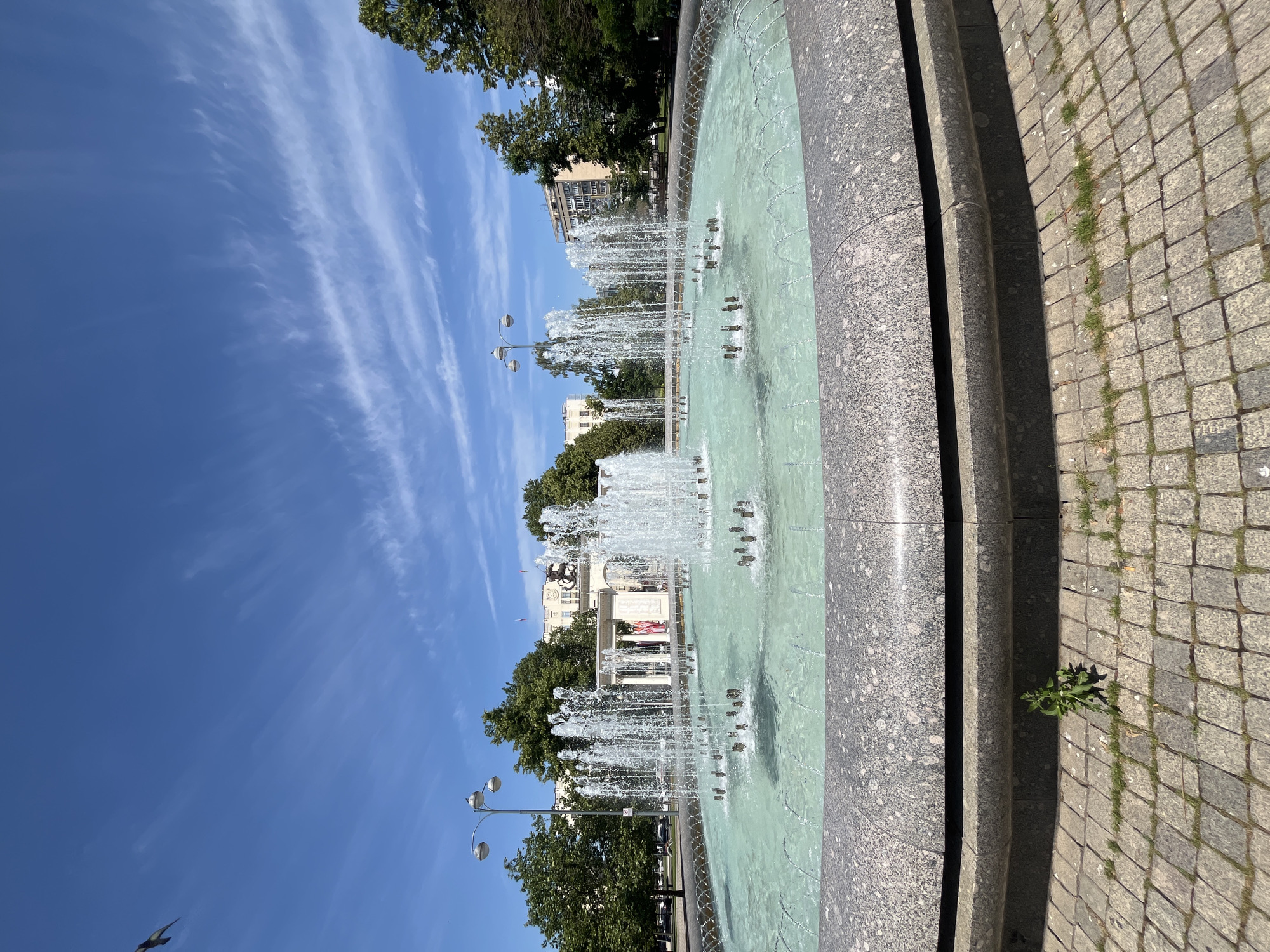 A wide-angle photograph captures a public fountain on a sunny day. The fountain is large and circular, filled with turquoise water that is actively being sprayed from multiple jets arranged in concentric circles. The fountain's edge is bordered by a thick band of granite with a speckled pattern. Outside the fountain, the ground is paved with interlocking cobblestones, with patches of grass and a small weed growing in the foreground.
In the background, several tall, leafy green trees line the perimeter of the area. Beyond the trees, there are buildings, including a large, light-colored building with columns and a statue or monument at its center. Streetlights with multiple lamps are visible among the trees. The sky is a clear, vibrant blue with thin, wispy white clouds. A bird is captured in flight in the upper left quadrant of the image. The overall impression is a well-maintained urban park or public square. The location is confirmed to be Krasnodar, Russia, specifically the Teatralnaya Square.