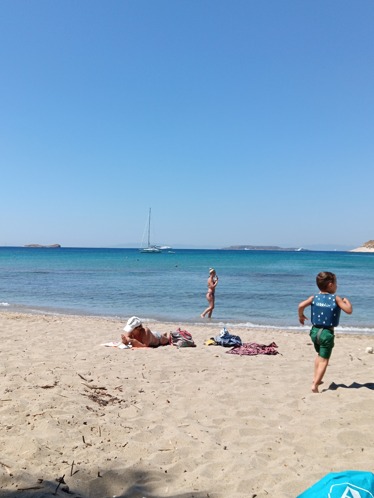 The image depicts a sunny day at a beach in Agia Marina, Greece. The scene is filled with people enjoying the clear blue sky and tranquil sea. In the foreground, a person lies on the sand wearing a white cap and appears to be reading. To the right, a young boy in a blue life vest and green shorts is running towards the sea. In the background, a woman in a bikini stands in the shallow water, possibly taking a photo. Further out in the ocean, a sailboat is anchored, with other distant boats and islands visible on the horizon. The water is a beautiful turquoise color, indicating a warm and pleasant day. The overall atmosphere is one of relaxation and enjoyment of a summer vacation. There is no visible text in the image.