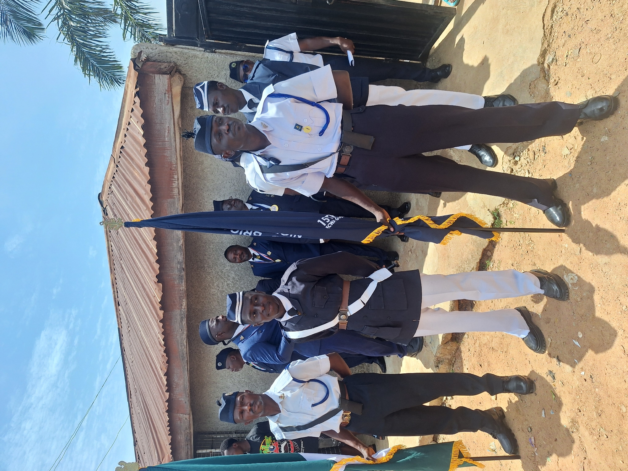 A group of men in uniforms are standing in a row. They are holding flags. The setting appears to be outdoors in Sabon Tasha, Nigeria. The location looks to be near a building with a corrugated metal roof, suggesting a more residential or less developed part of the city. The uniforms worn by the men feature different color schemes with white, navy blue, and black elements. The uniforms include caps and the men are wearing polished black shoes.