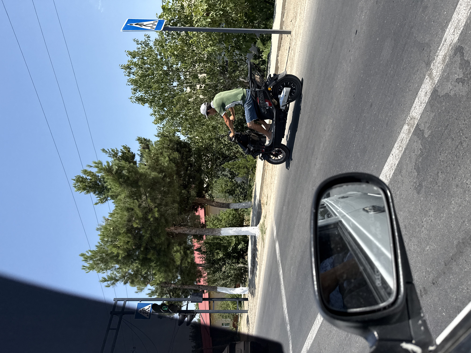 A person wearing a green shirt, denim shorts, and a white baseball cap is riding a black electric scooter on a paved road. The scooter has the word "CHEETAH" on its side. The road has a white painted line down the center. To the left of the road, a traffic light is visible, displaying a green signal. A pedestrian crossing sign is posted to the right of the road. Trees line the side of the road, and some have their lower trunks painted white. In the foreground, a car's side mirror reflects a portion of the car's interior and the road. The sky is clear and blue. The scene appears to be a street in the city of Kahramon, Uzbekistan.