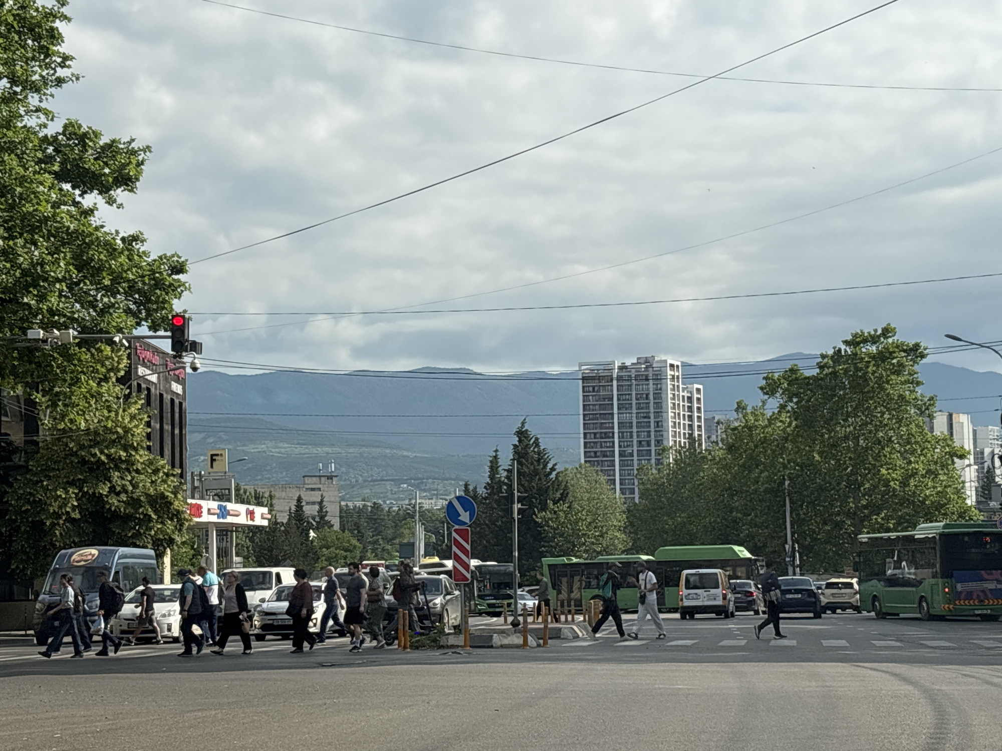 The image depicts a street intersection in Tbilisi, Georgia, under a cloudy sky. In the foreground, several people are crossing a busy road at a pedestrian crossing. Vehicles, including cars and buses, are visible on the road. On the left side of the image, a traffic light is showing red. Trees and buildings, including a tall apartment complex, are present in the background, with mountains visible in the distance. The overall scene suggests a typical urban environment with active traffic and pedestrian movement.