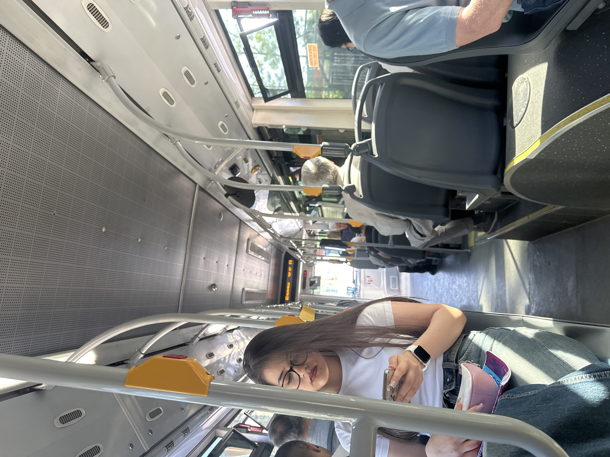 The image shows the interior of a public bus. The bus is filled with several passengers, including a woman with long dark hair, wearing glasses, a white t-shirt, jeans, and an Apple watch. She is sitting down and looking at her phone. The scene includes gray seats, metal bars, yellow stop buttons, and the bus's ceiling with vents. The presence of signage in Cyrillic script suggests the location is likely Sofia, Bulgaria, as this is the official alphabet used in the country.