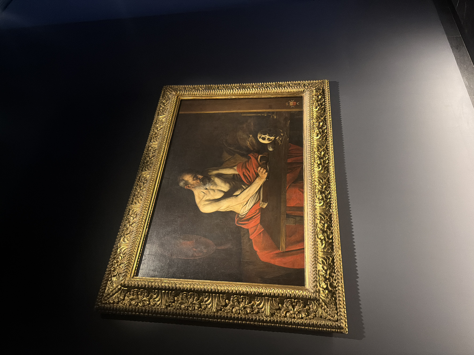 The image depicts the painting "The Martyrdom of Saint Peter" by Caravaggio, hanging on a wall. The painting shows Saint Peter being crucified upside down. The artwork is framed in an ornate, gold frame. The painting is located in the city of Valletta, Malta. The wall around the painting is grey, with lighting that casts shadows on the wall.