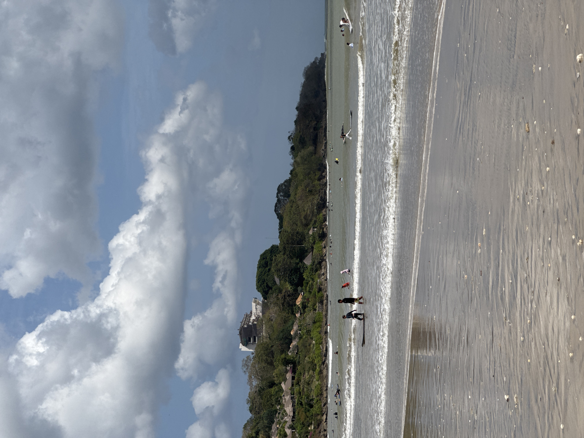 This image depicts a beach scene in Kuta Selatan, Indonesia, on a partly cloudy day. The foreground features a sandy beach with wet sand reflecting the sky and clouds. Small shells are scattered across the sand. Gentle waves are breaking near the shore. In the middle ground, the ocean is populated with numerous surfers on their boards. Some are riding waves, while others are paddling or waiting. Two young boys are walking in the shallow water, one of them with a surfboard. In the background, a lush green hillside rises, dotted with small thatched-roof structures and a larger, modern building on the crest of the hill. The sky is a mixture of blue sky and dramatic, puffy white and gray clouds.
