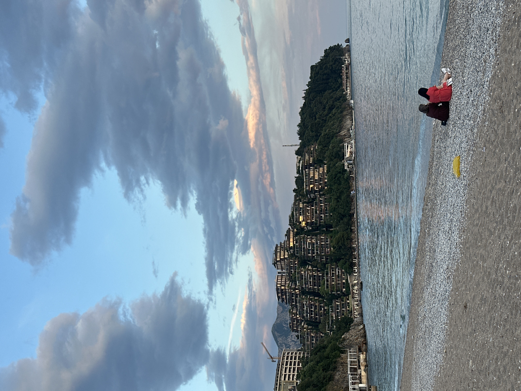 The image shows a shoreline scene in Budva, Montenegro at dusk or dawn.
Environment: The scene is dominated by a coastline with buildings built into a steep, densely vegetated hillside. The sky is filled with gray and blue clouds, and the sea is calm. A pebble beach lines the shore. Construction is visible in the lower left.
People: Two people are sitting on the beach, positioned close to the water's edge. One is wearing a red garment. The other person is wearing brown.
Objects: Buildings, trees, sea, beach, pebbles, construction cranes, and clouds. A yellow object is on the beach near the sitting people.