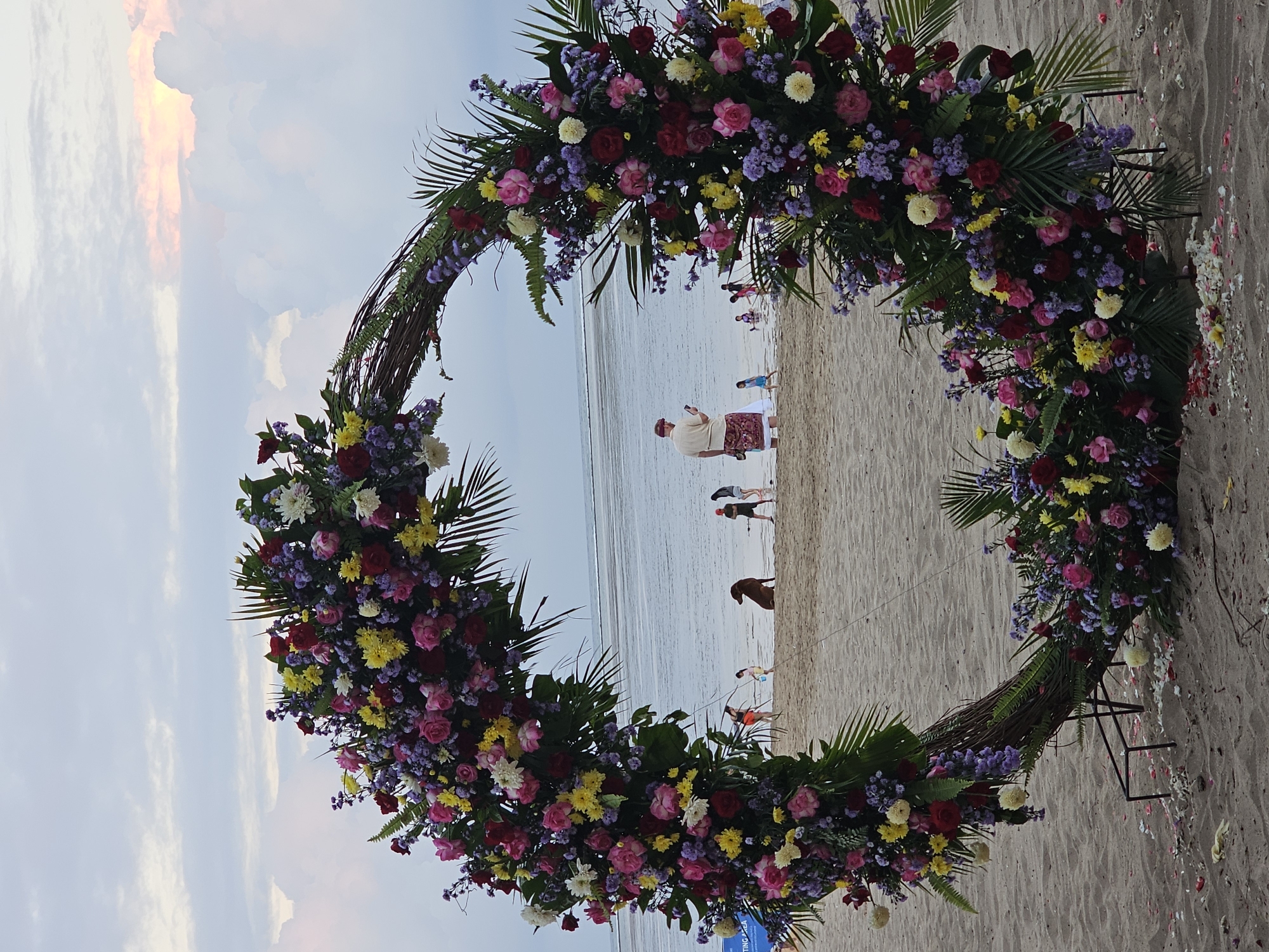 A large, circular floral arch, decorated with an assortment of pink, red, yellow, and white flowers, as well as greenery, stands on a sandy beach. The arch is made of woven branches and is supported by a dark metal frame. In the background, the ocean is visible, with a calm horizon. Several people and a dog are scattered across the beach, some walking near the water, others standing. The sky above is partly cloudy with hints of orange and pink, suggesting either sunrise or sunset. A man in a light-colored shirt and patterned skirt stands facing away from the camera, looking at his phone. The overall scene suggests a picturesque outdoor event, possibly a wedding or photoshoot, set against a coastal backdrop.