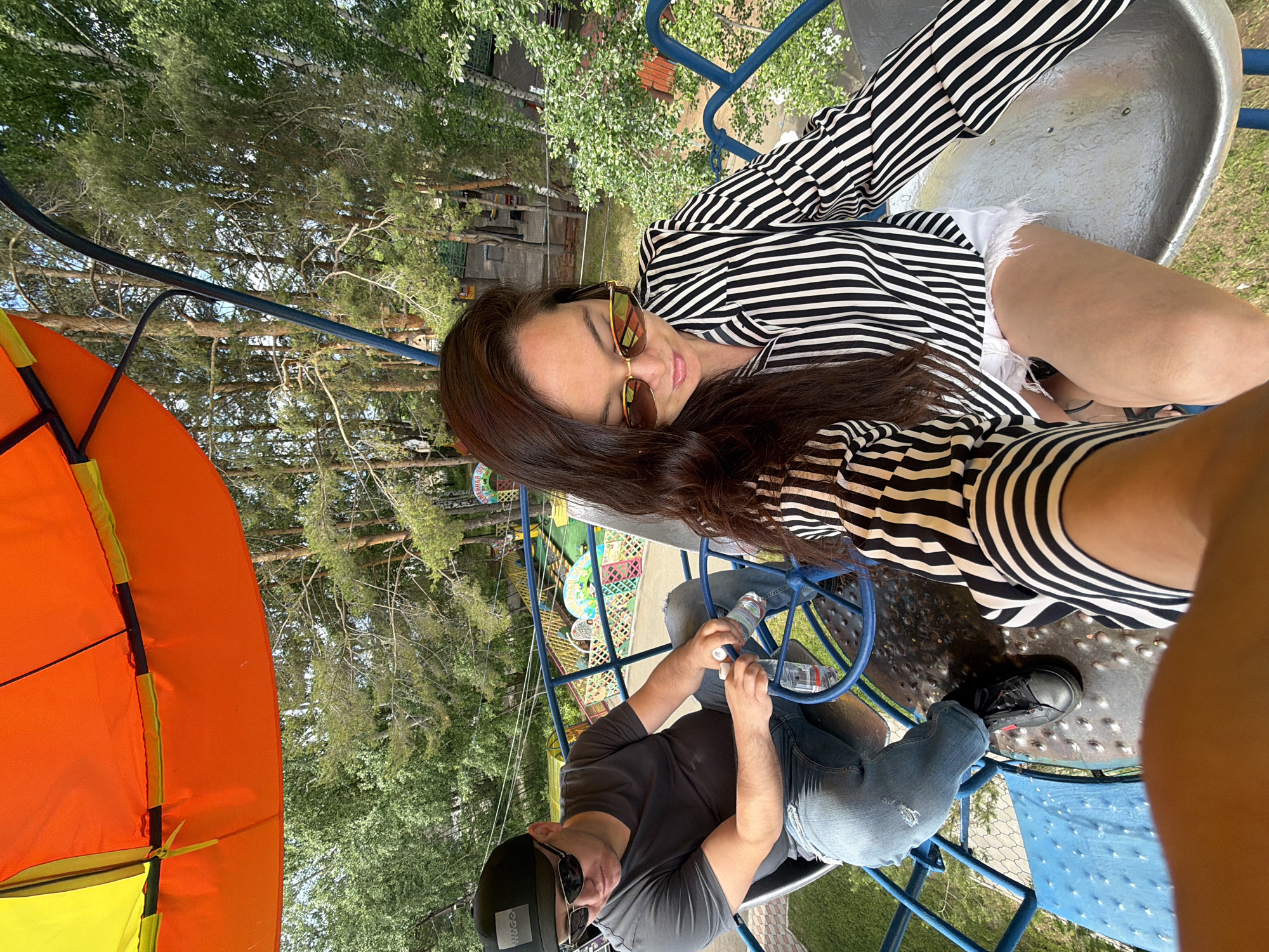 An outdoor, high-angle shot captures two individuals on an amusement park ride during what appears to be a sunny day. The image is rotated counter-clockwise.
A woman is positioned prominently in the upper right, wearing sunglasses, a black and white striped long-sleeved top, and white shorts. She has long, dark hair flowing over her shoulder and an expression of calm relaxation. Her lower body and legs are visible on the ride's textured gray platform.
Seated opposite her, in the lower center, is a man. He wears a dark t-shirt, blue jeans, and sunglasses. A black baseball cap on his head features the text "HUGO". He is holding two plastic water bottles. A blue steering wheel-like handle is in front of him on the ride.
The ride itself appears to be a multi-person spinning or rotating attraction, characterized by blue metal bars and a gray, metallic floor. To the left, a vibrant orange and yellow canopy or tent structure is partially visible. The background reveals a lush, green environment with numerous trees, including what appear to be birch and pine trees. Other colorful amusement park rides and structures, some with visible Cyrillic text that is largely illegible, can be seen in the distance, indicating a recreational area in Chaikovsky, Russia. The overall atmosphere suggests a leisurely and enjoyable outing.
