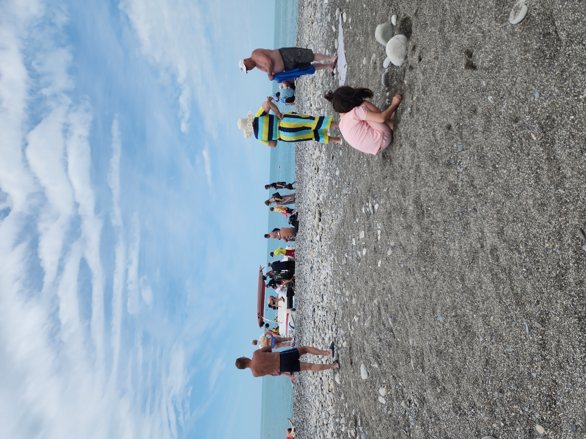 The image captures a scene at a pebble beach in Sochi, Russia, with a partly cloudy sky. The beach is crowded with people engaging in various activities. Some individuals are standing near a small motorboat, while others are swimming in the water. In the foreground, a woman in a striped dress and a sun hat stands next to a seated child wearing pink. Near them, a man is toweling off. Another man is further down the beach.