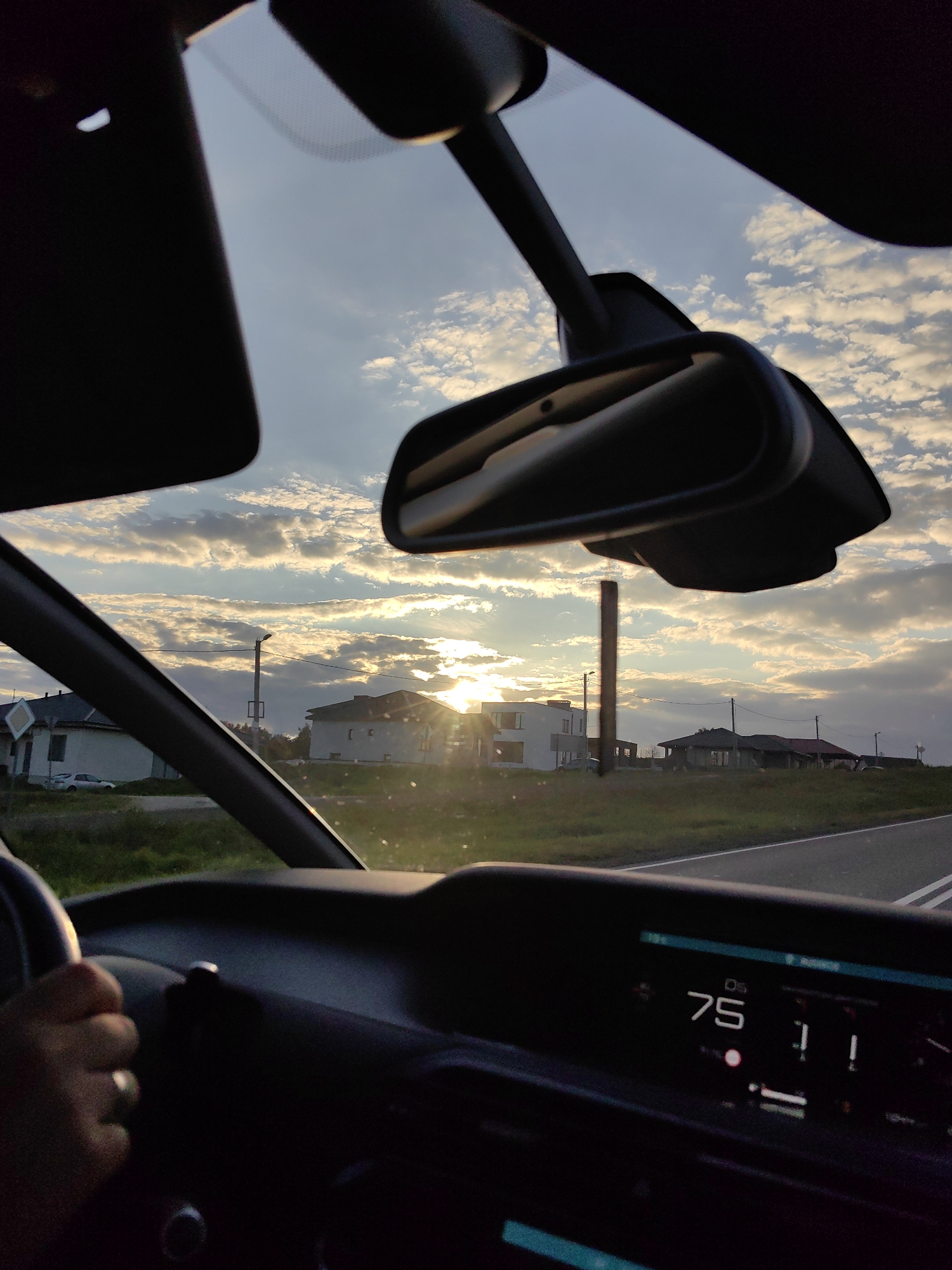 The image is taken from inside a car, looking out the windshield towards a sunset. The sun is low on the horizon, casting a warm glow through scattered clouds. In the foreground, the car's dashboard is visible, with a digital display showing "75" and some icons. A hand is on the steering wheel, with a ring on the finger. Outside, there are houses and a tall pole with wires. The overall setting suggests a rural or suburban area at dusk. Based on the available information, the location is identified as Dzyarzhynsk, Belarus.
